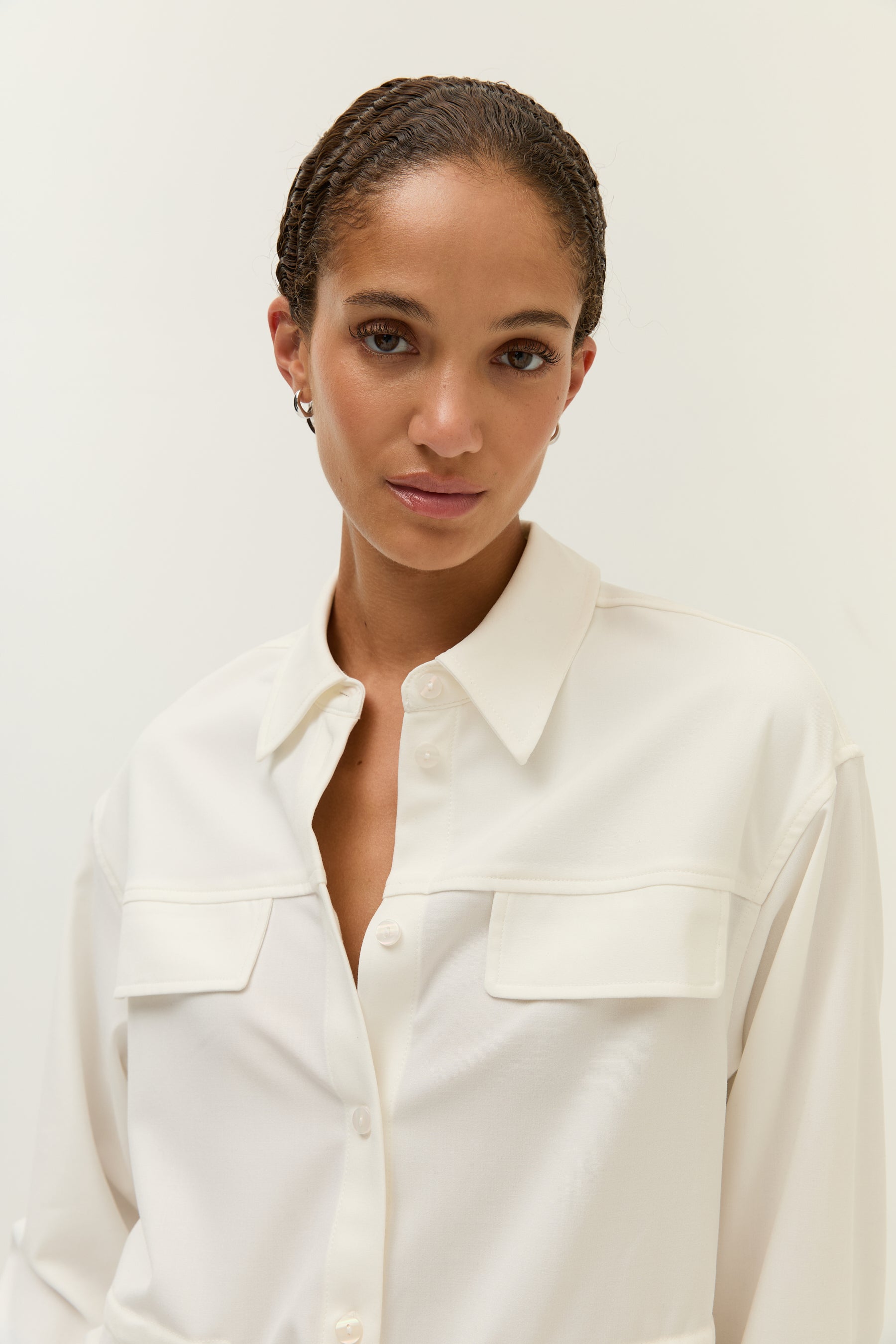 Frau in entspannter Pose mit geöffnetem weißen Hemdblusenshirt, kombiniert mit brauner, weit geschnittener Hose.