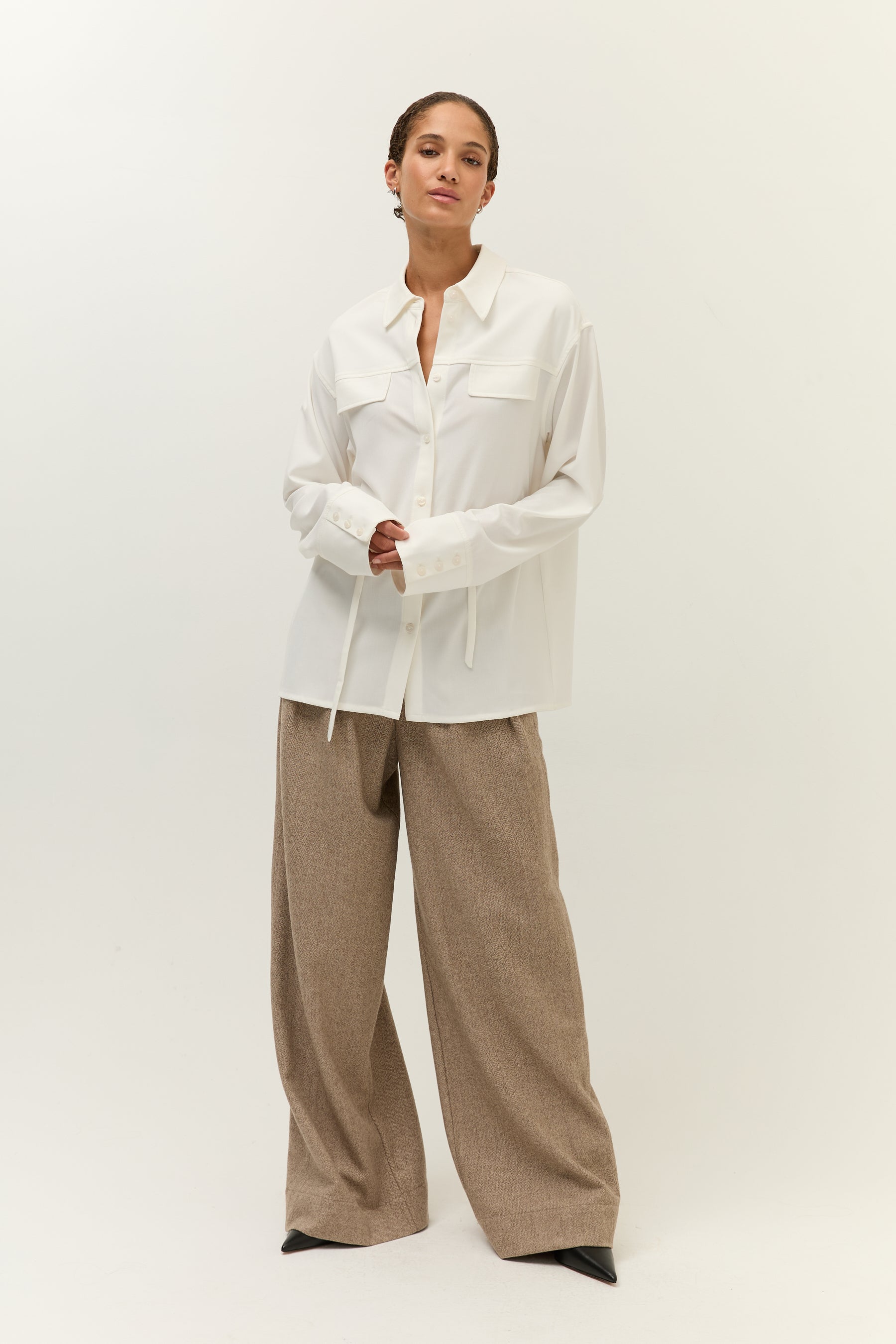 Ganzkörperaufnahme der Frau mit geöffnetem weißen Hemdblusenshirt, darunter sichtbares Dekolleté, kombiniert mit brauner Marlenehose und spitzen schwarzen Schuhen.