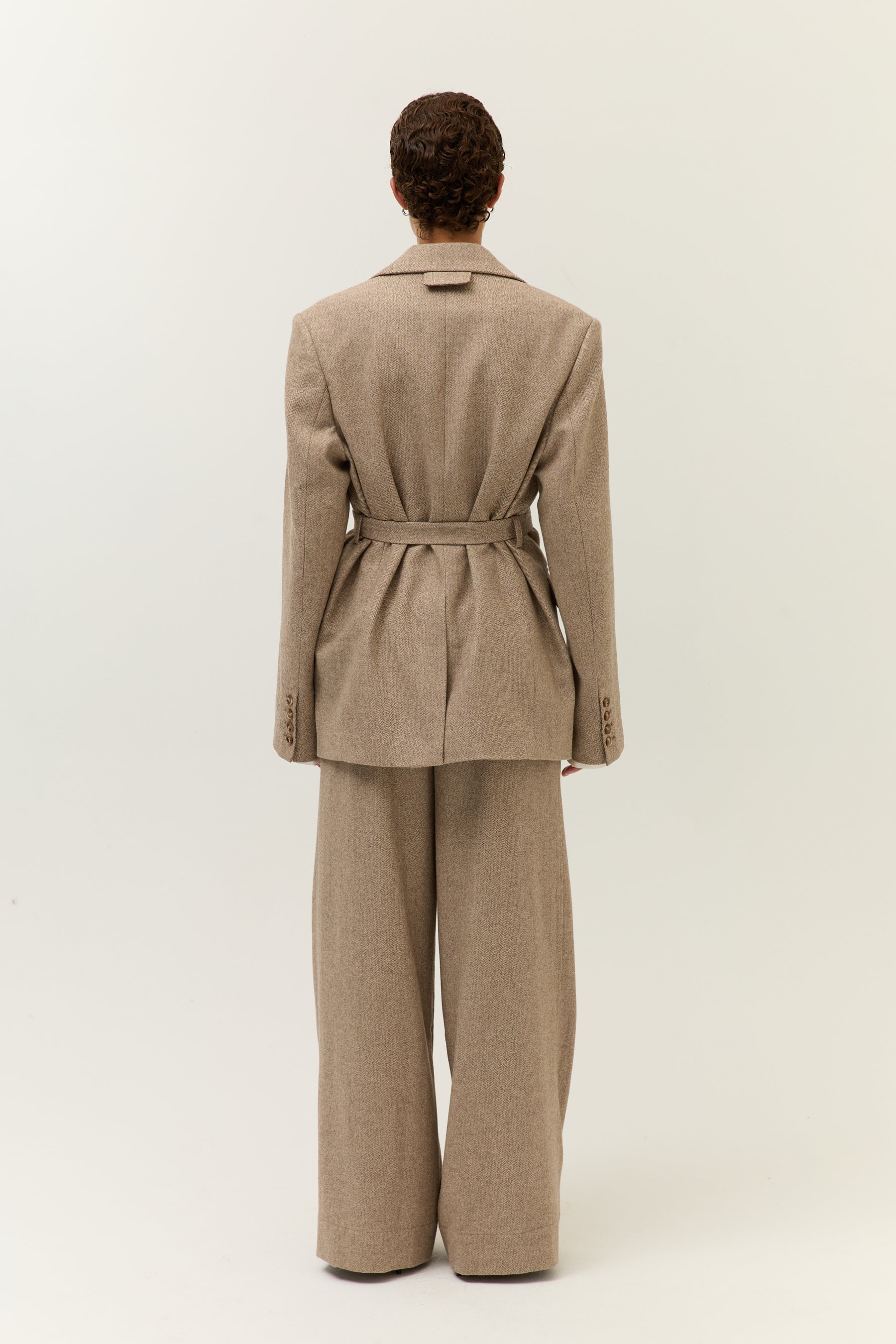 Ganzkörperaufnahme des Models von hinten, der Blazer ist mit Gürtel auf Taille gebunden und fällt über die weite Hose.