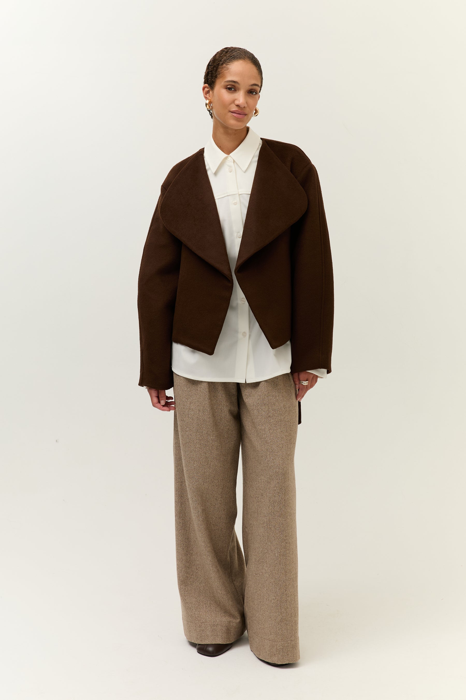 Vollansicht frontal, offen getragen: Frau in einer dunkelbraunen Jacke mit großem Revers über einer weißen Hemdbluse und weit geschnittener beigefarbener Wollhose.
