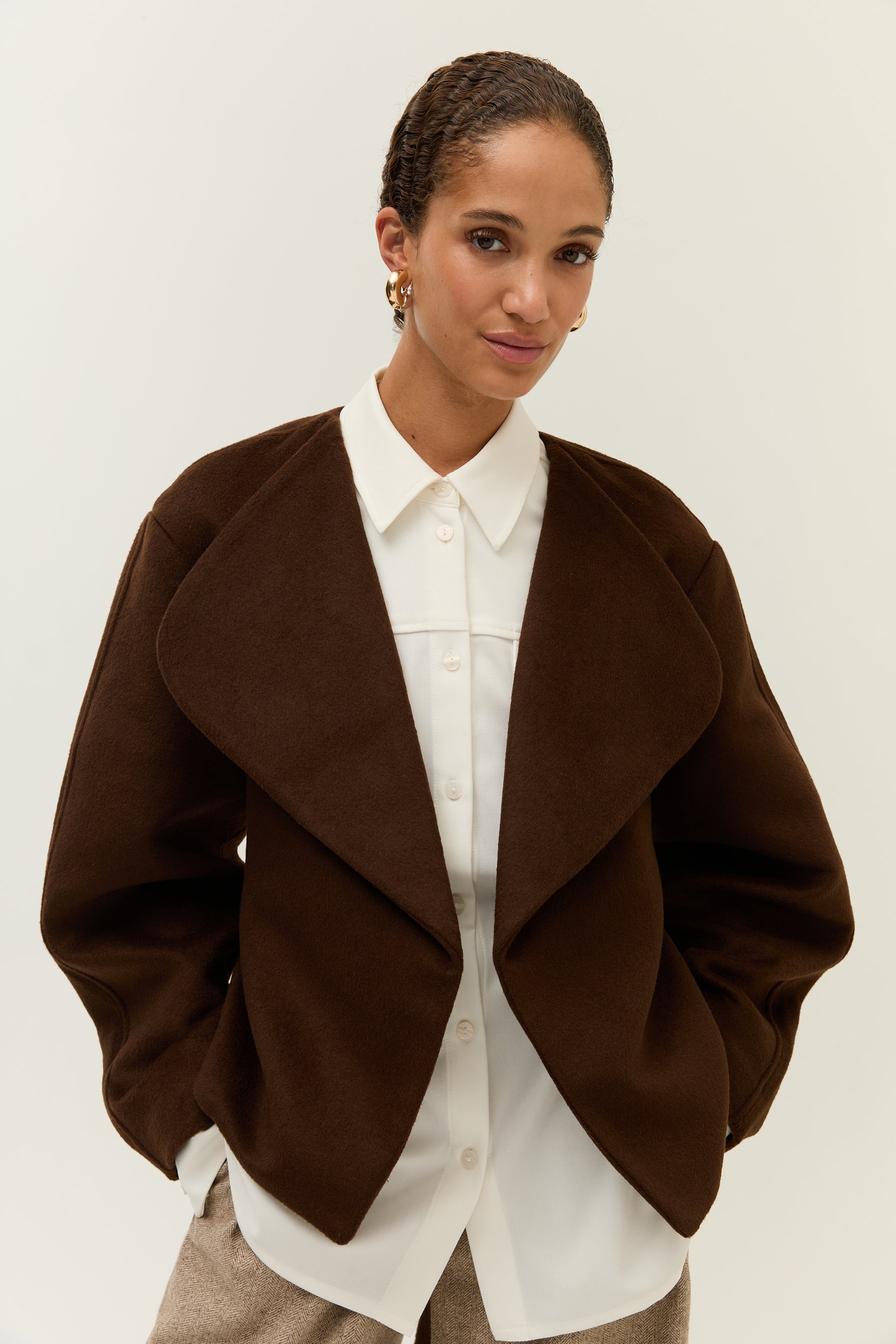 Frau mit zusammengebundenem Haar trägt eine elegante, kastig geschnittene Jacke in Dunkelbraun mit betont breitem Revers. Darunter eine weiße Hemdbluse mit klassischem Kragen und sichtbarer Knopfleiste. Sie blickt leicht zur Seite und lächelt dezent. Der Hintergrund ist neutral und cremefarben,