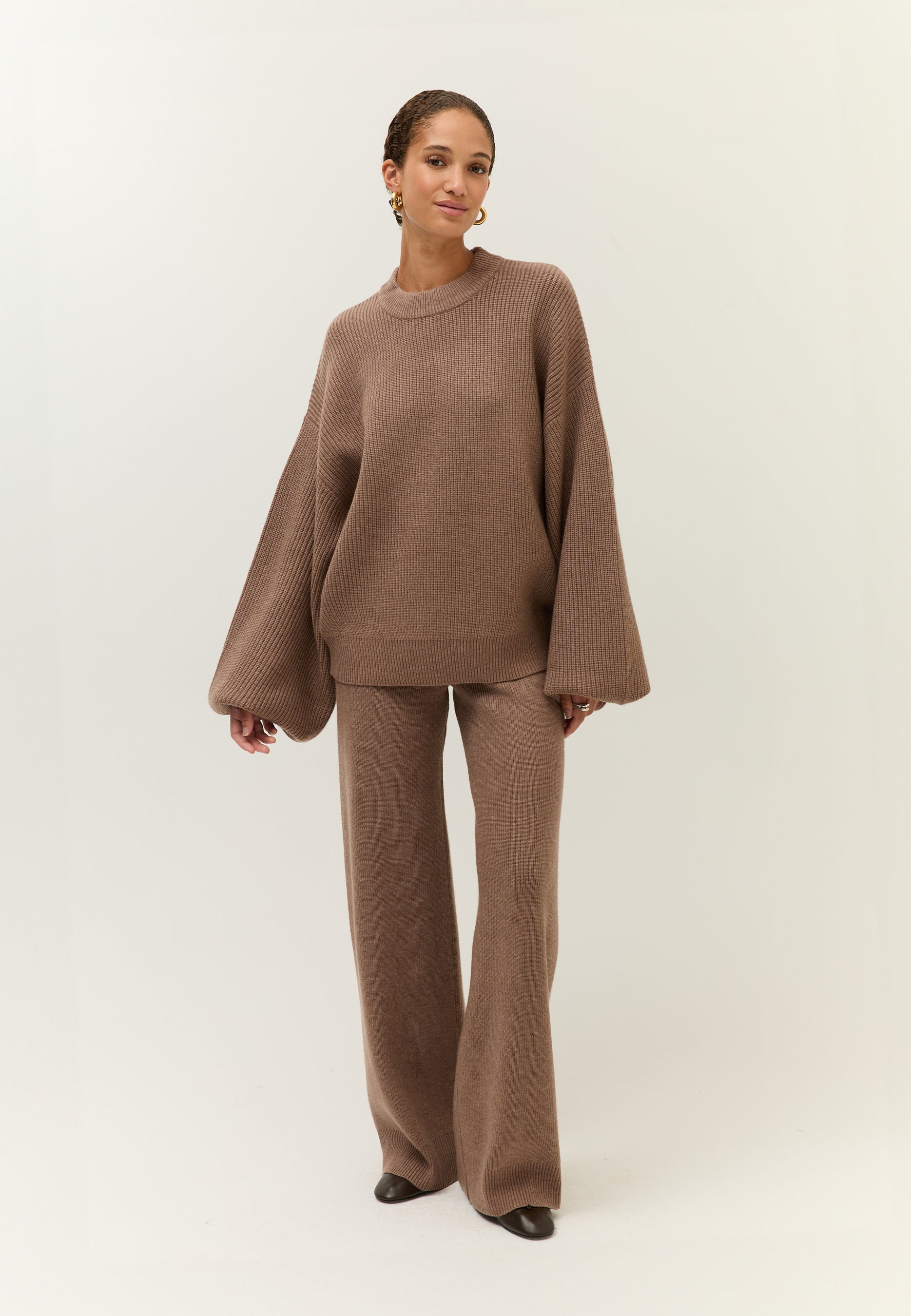 Bild 2: Ganzkörperansicht der Frau in einem braunen Strick-Ensemble mit weitem Pullover und passender Hose. Sie steht leicht seitlich und schaut ruhig nach vorn.