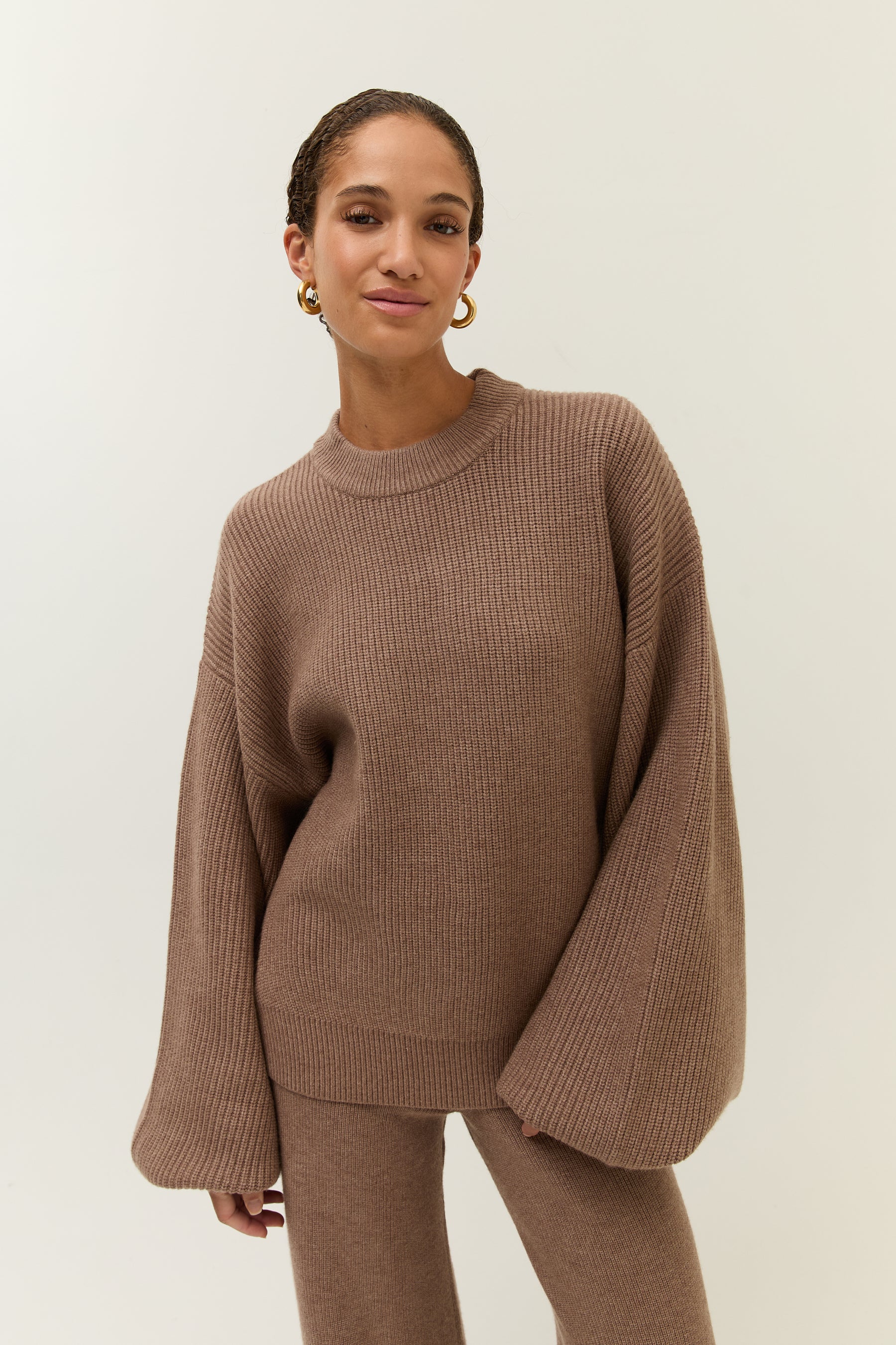 Bild 1: Frau trägt einen locker sitzenden, gerippten Strickpullover in warmem Braunton mit weiten Ärmeln. Sie blickt leicht lächelnd in die Kamera, goldene Creolen schmücken ihre Ohren.