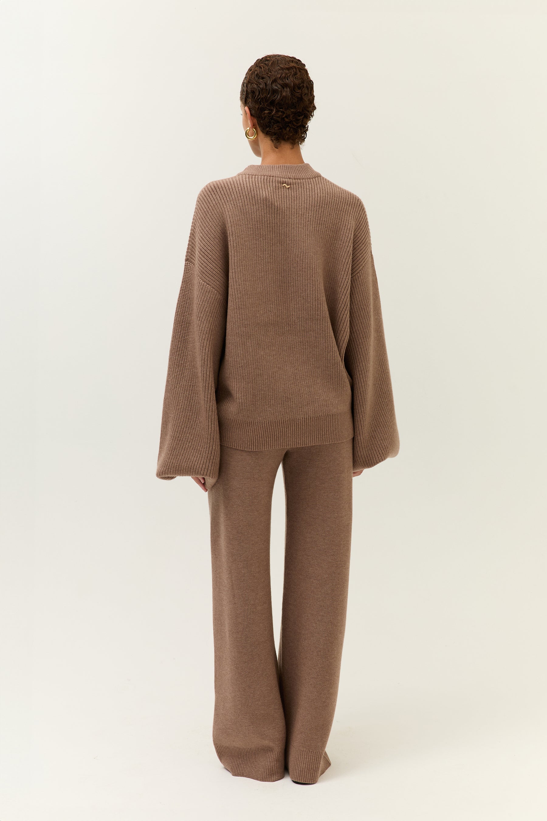 ückansicht des Modells, das den Pullover mit einer Hand locker anhebt. Der Schnitt der Hose ist betont figurumschmeichelnd.