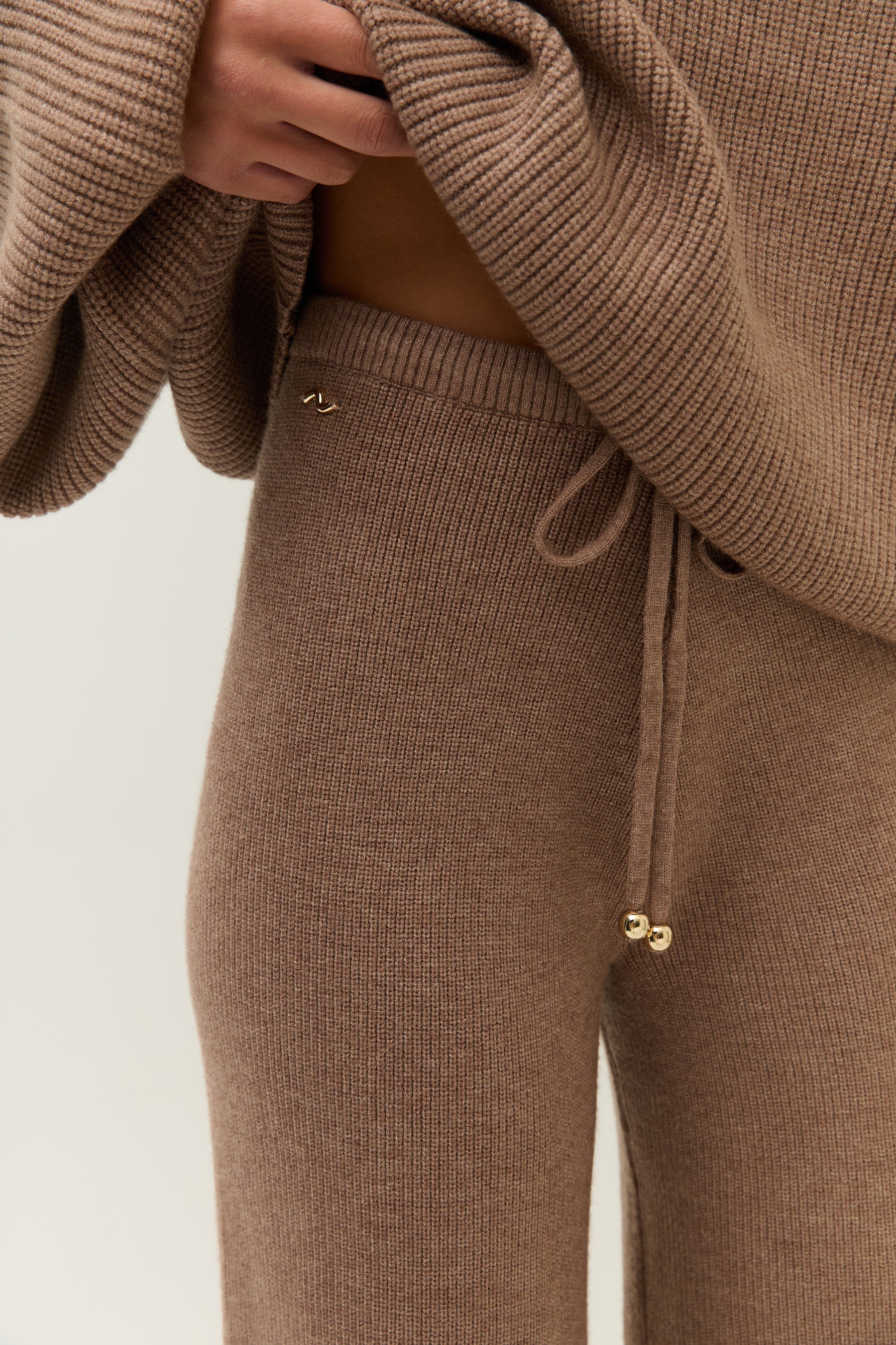 Nahaufnahme der Hose auf Taillenhöhe. Sichtbar sind der elastische Bund, der Kordelzug mit goldenen Endstücken und das gewebte Lezu-Logo in Gold.