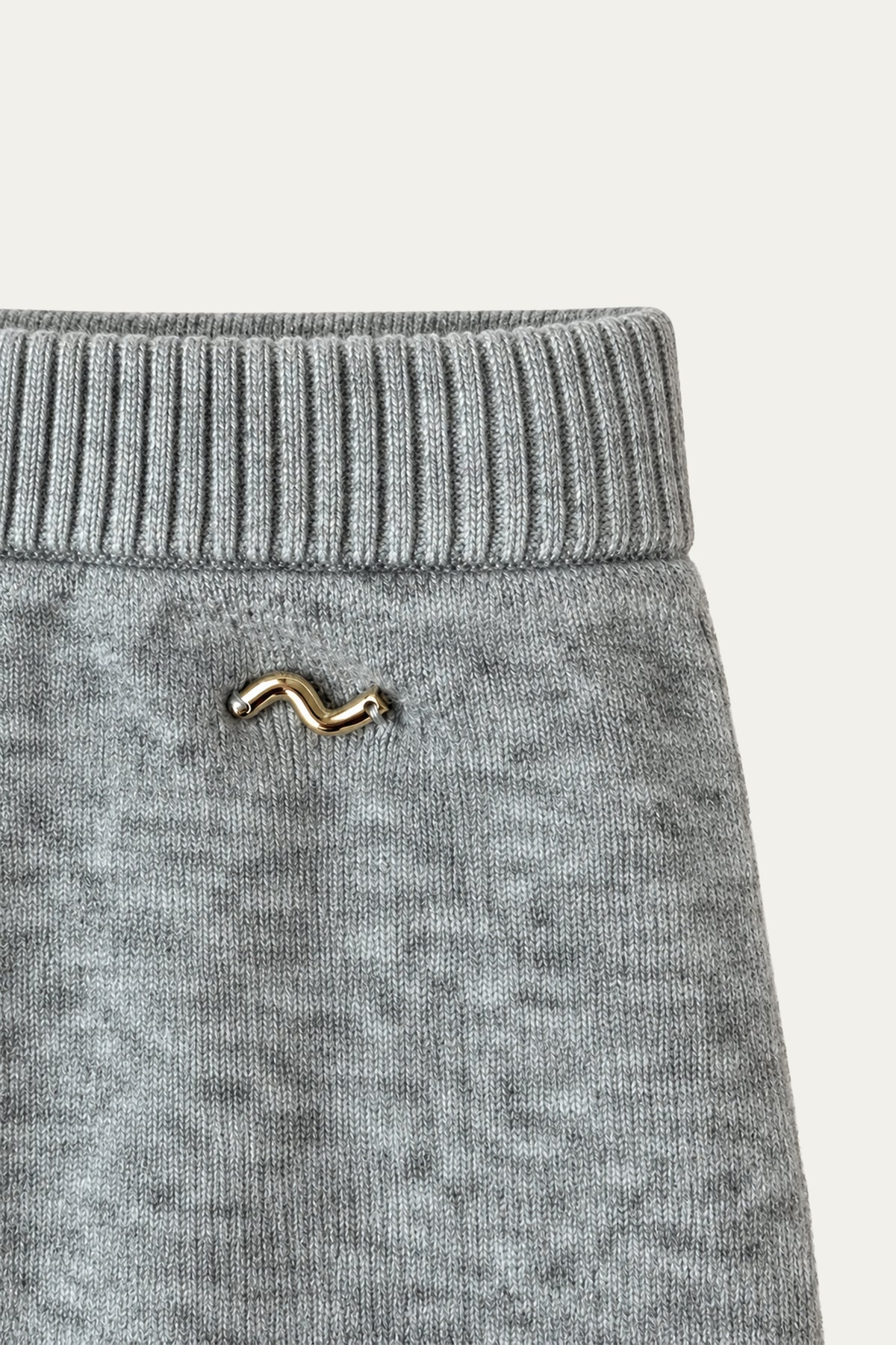 Detailansicht einer hellgrau melierten Strickhose mit breitem Rippstrickbund und kleinem goldfarbenem Logo-Emblem.