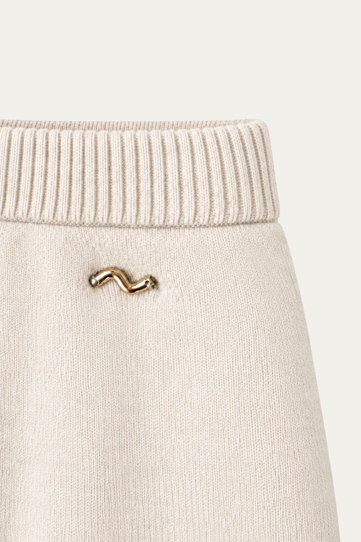 Detailaufnahme einer cremefarbenen Strickhose mit breitem geripptem Bund und kleinem goldfarbenem Logo.