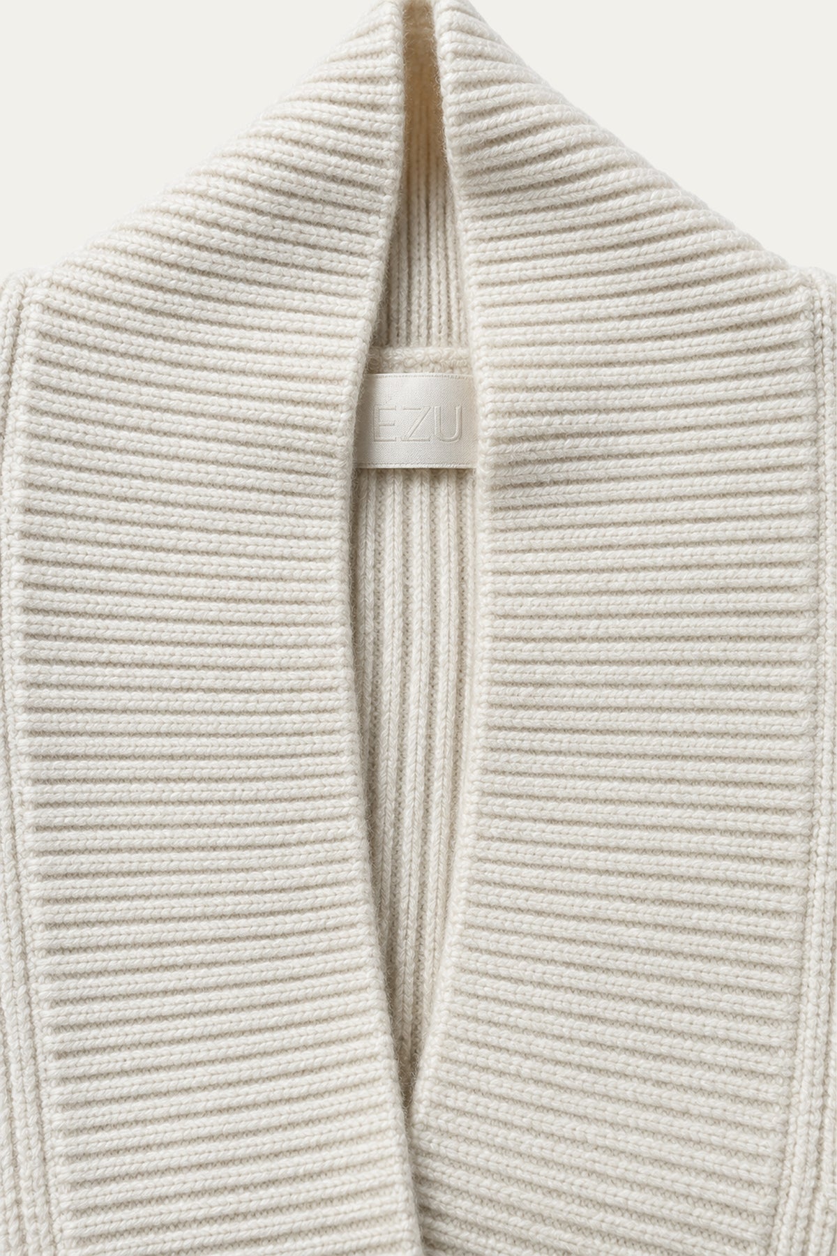 Detailaufnahme eines cremefarbenen Strickoberteils mit breitem geripptem Kragen und hellem LEZU-Label.