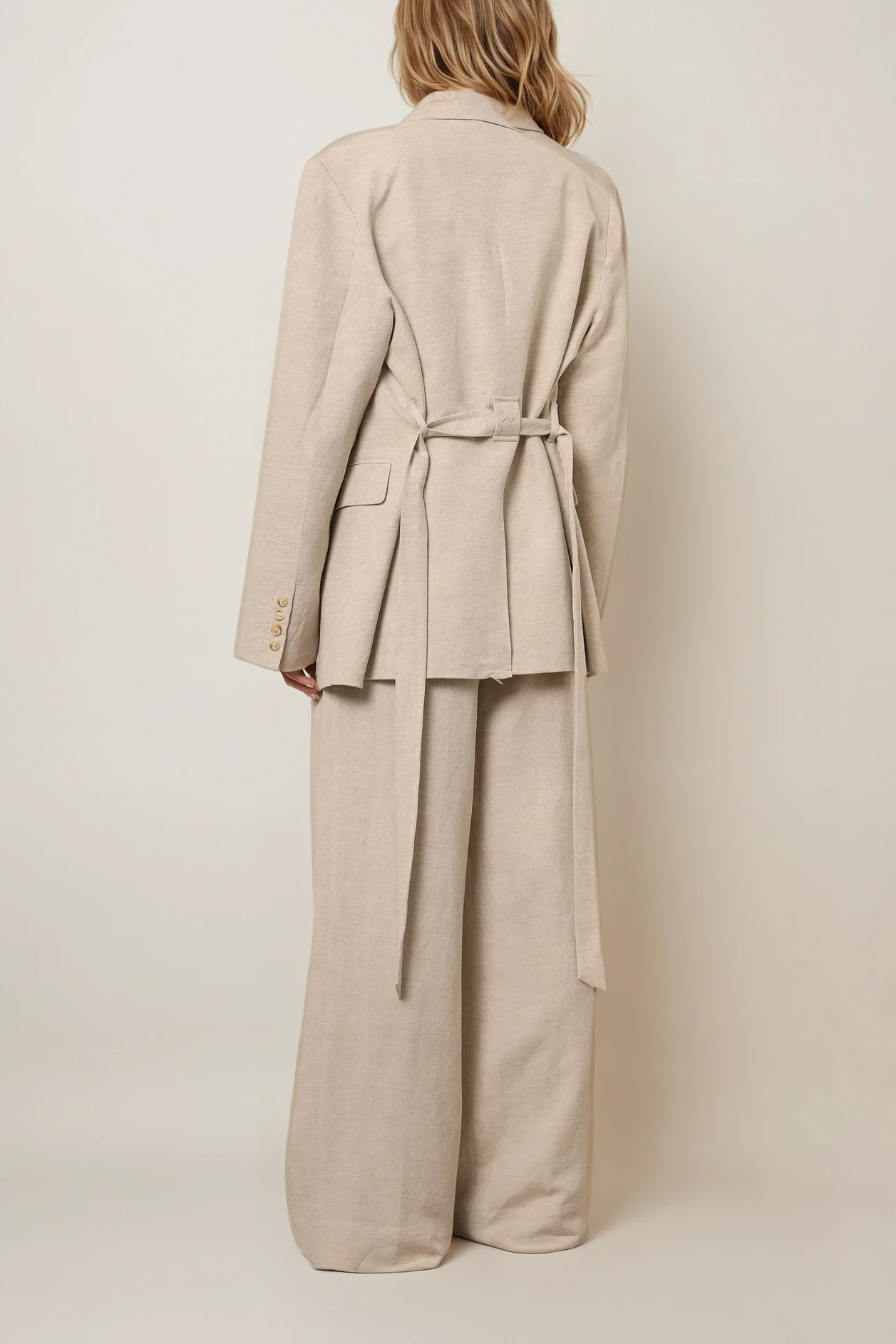 Rückansicht: Blazer in Warmgrau mit Taillengürtel und gerader Passform, kombiniert mit weiter Hose im gleichen Material.