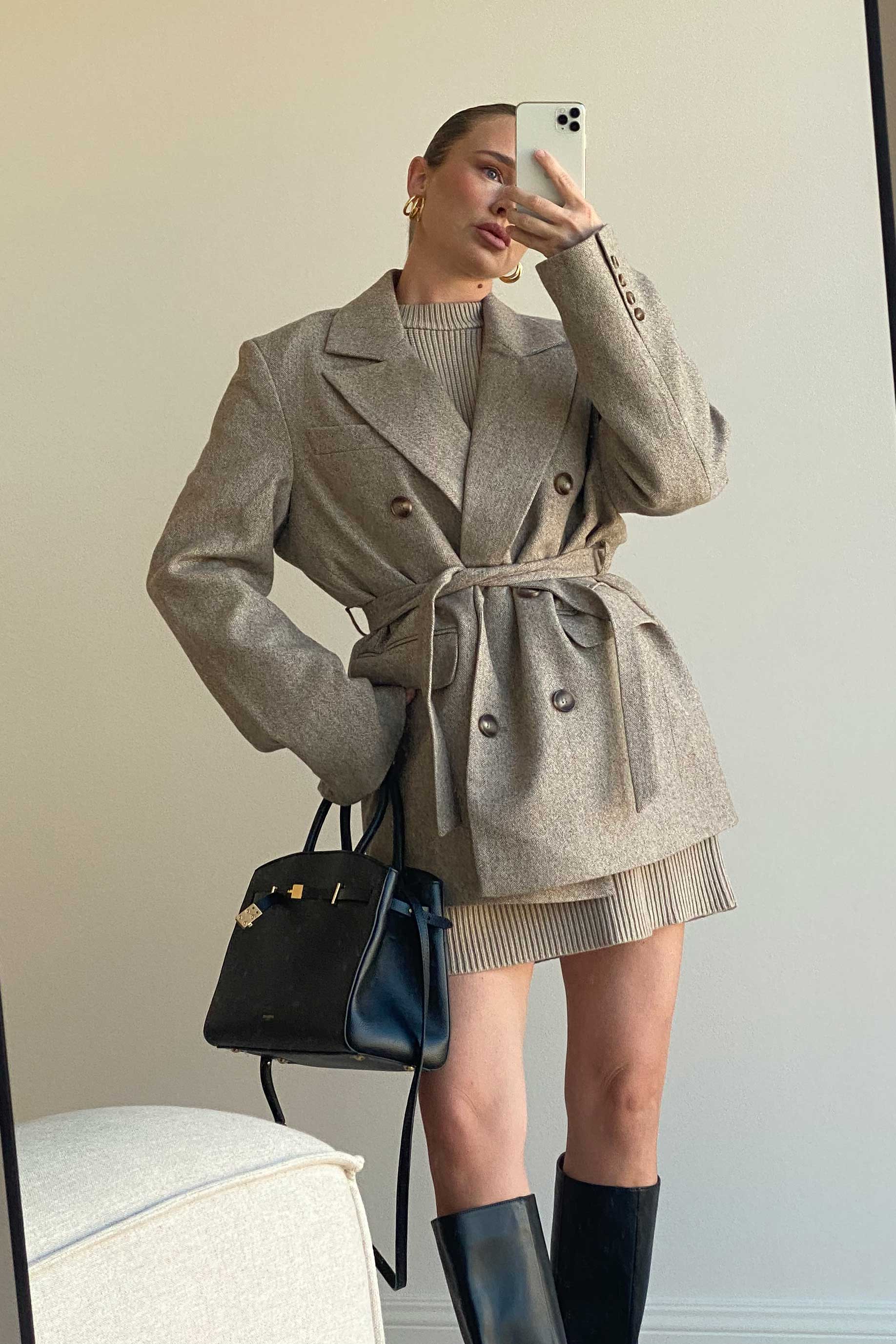 Spiegelselfie einer Frau in einem eleganten, beigefarbenen Oversize-Blazer mit Taillengürtel über einem gerippten Strickkleid. Sie trägt große goldene Creolen, hält eine schwarze Designer-Handtasche und kombiniert den Look mit kniehohen schwarzen Lederstiefeln. Das Outfit wirkt herbstlich und sophisticated.