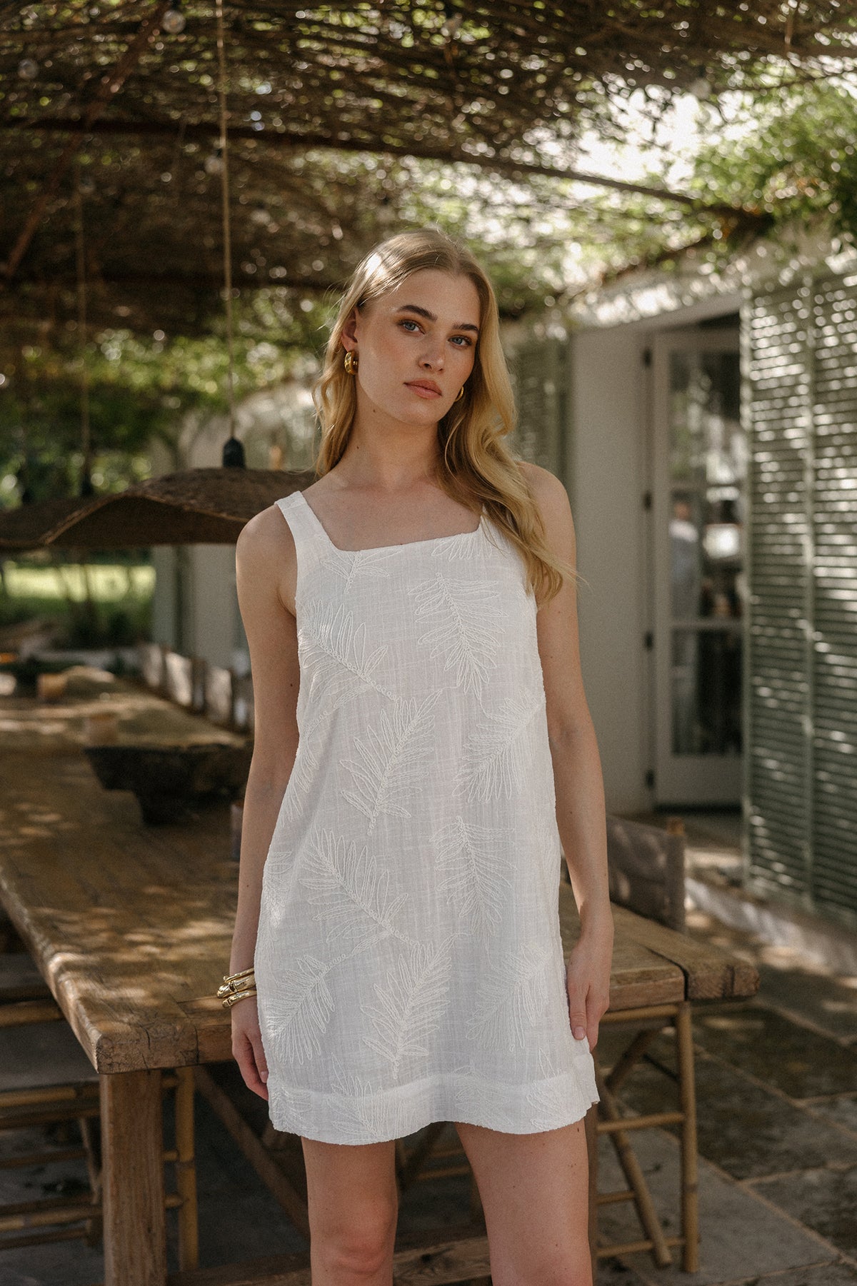 Model steht im Außenbereich unter einer Pergola und trägt ein kurzes, ärmelloses weißes Kleid mit breiten Trägern und dezentem Strukturmuster, kombiniert mit goldenen Armreifen