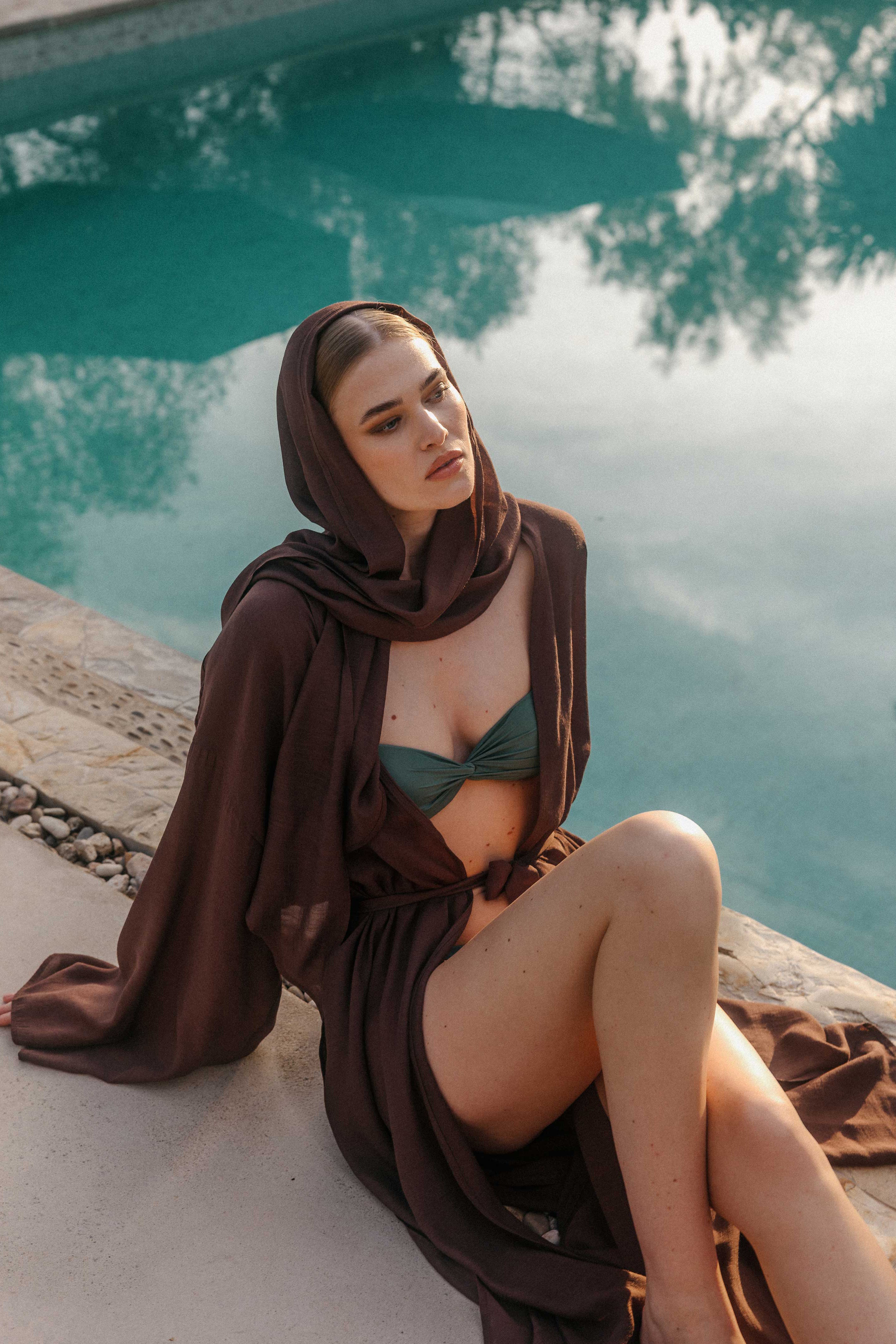 Model sitzt am Pool mit dunkelbraunem transparentem Überwurf und passendem Pareo, kombiniert mit einem grünen Bandeau-Bikinioberteil und locker drapiertem Schal.