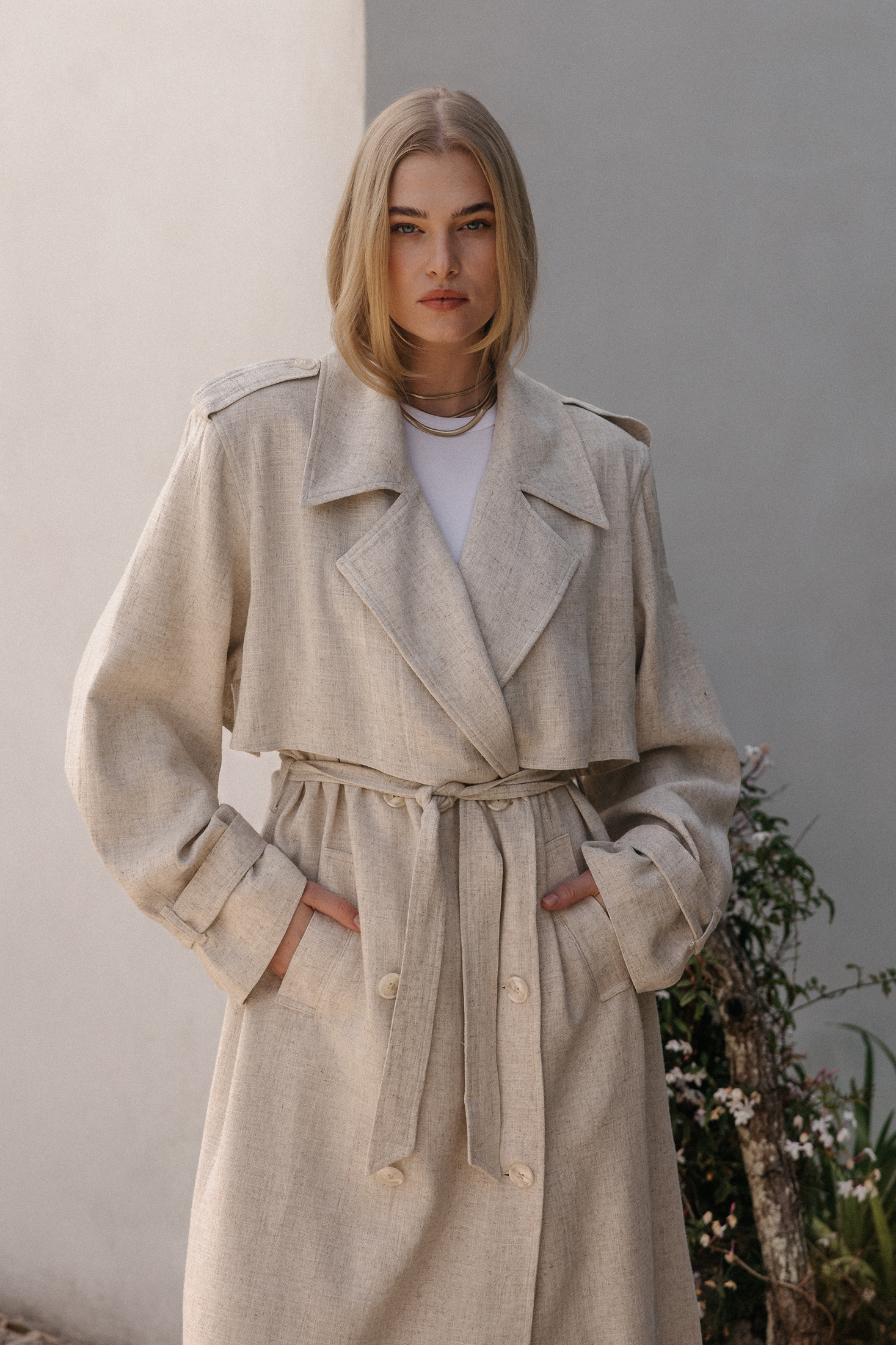 Model trägt einen langen beigefarbenen Trenchcoat mit Bindegürtel, doppelter Knopfleiste und weitem Kragen über einem weißen Oberteil, kombiniert mit goldfarbenem Halsschmuck