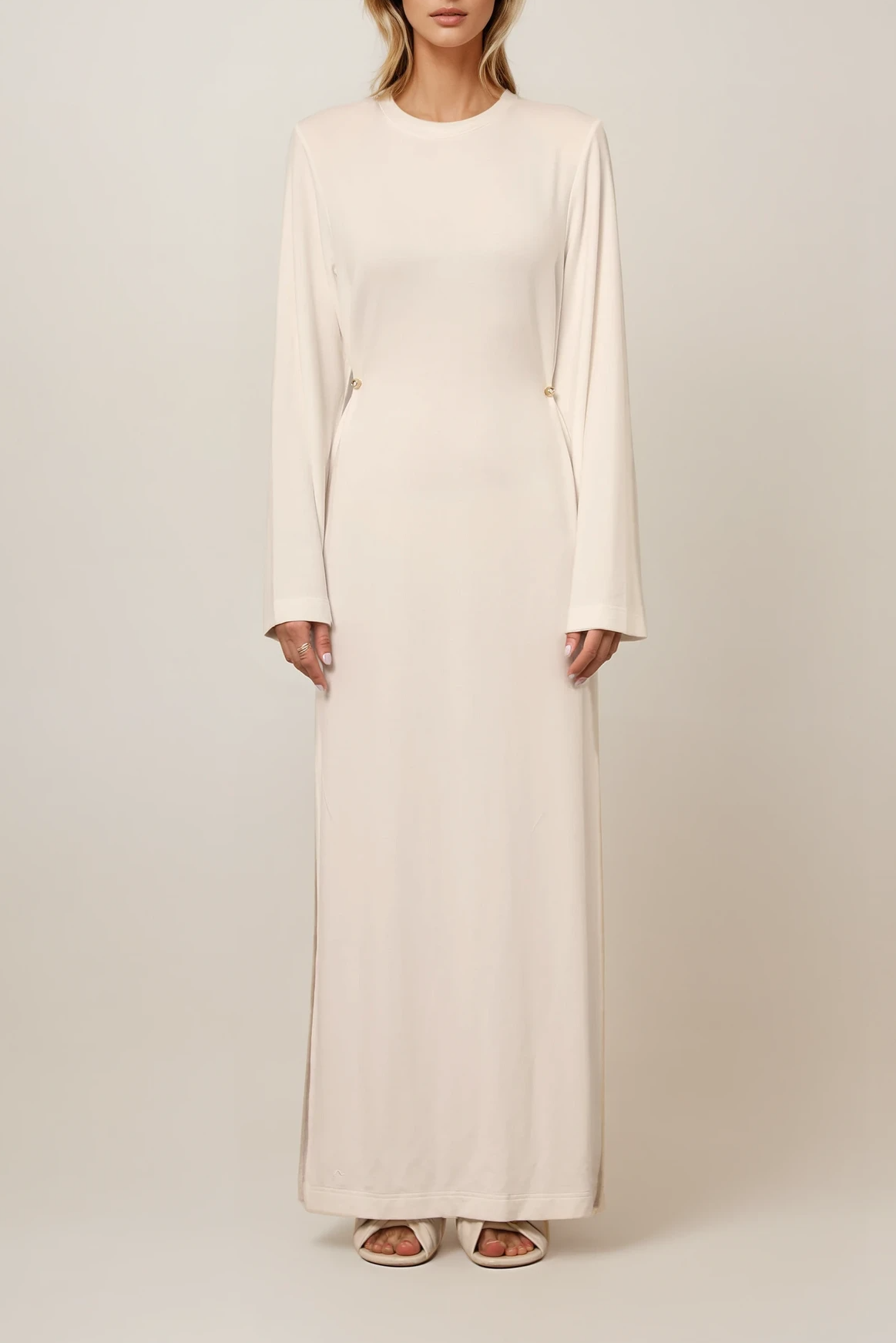Model trägt ein langes, fließendes Kleid in Off-White mit Rundhalsausschnitt und taillierender Passform durch seitliche Details.