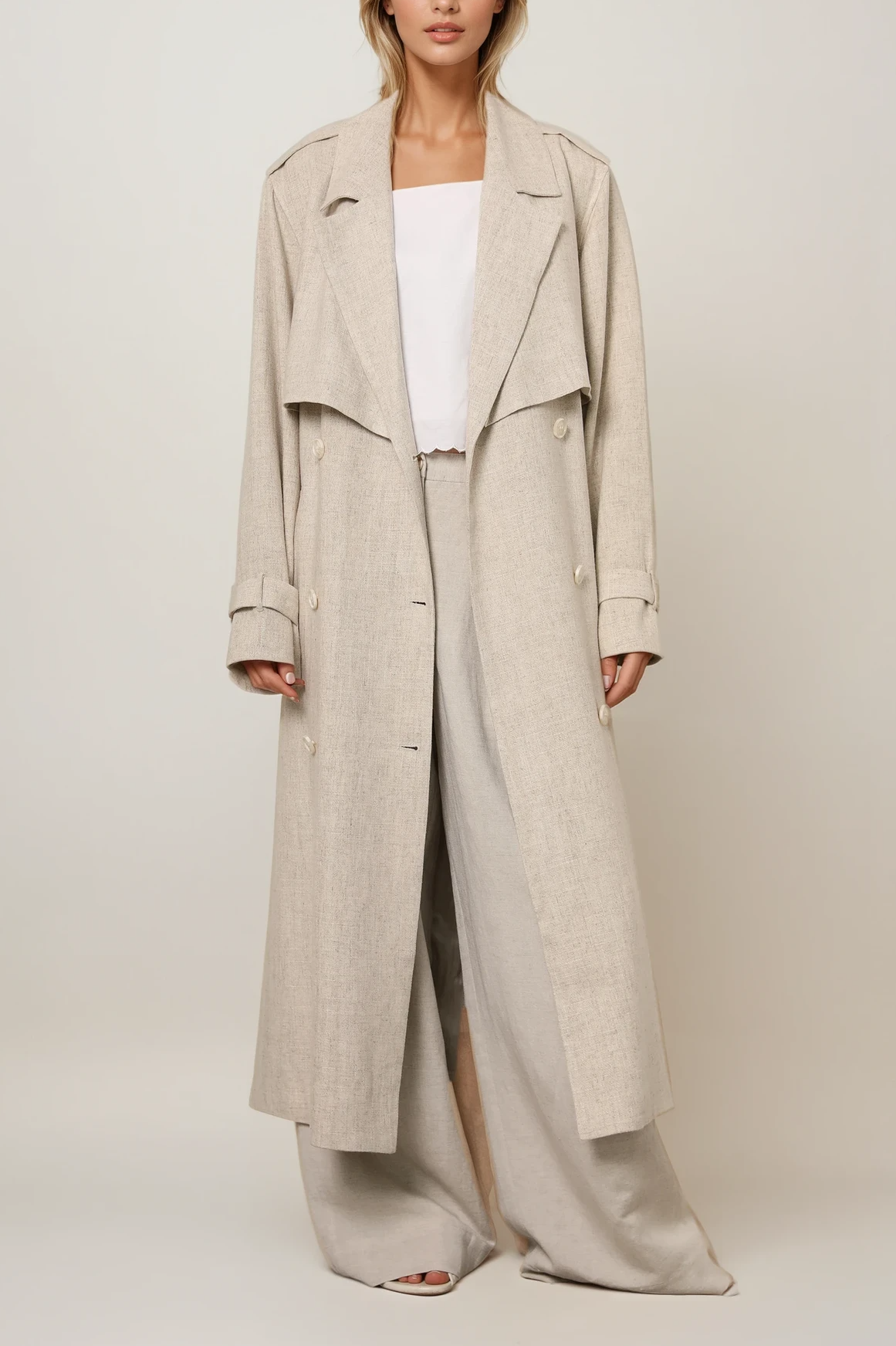 Model trägt einen langen Trenchcoat in Beige Melange mit Reverskragen und Bindegürtel, kombiniert mit weiter Hose.