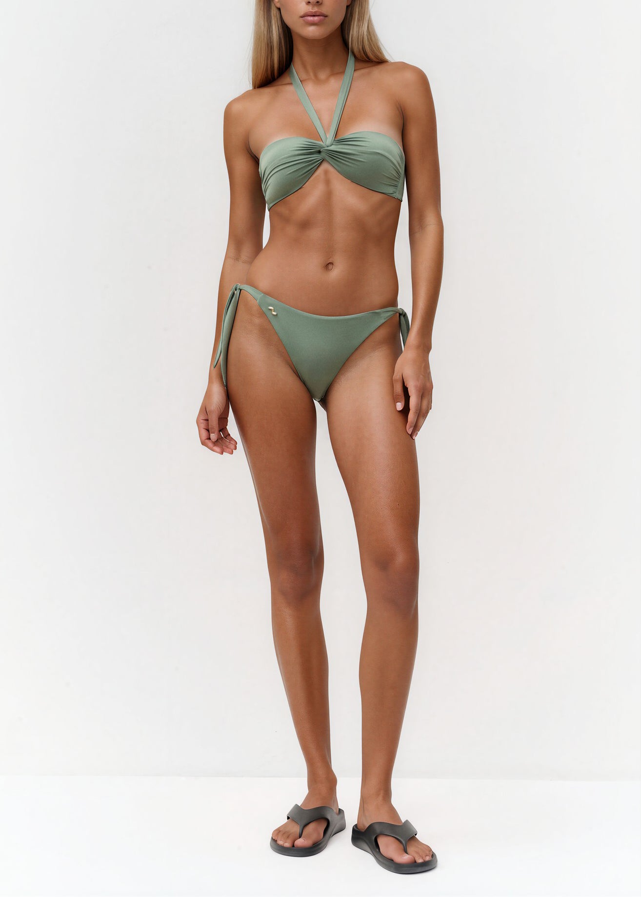 Model trägt einen olivgrünen Bandeau-Bikini mit gerafftem Oberteil und seitlich gebundener Bikinihose vor hellem Hintergrund.