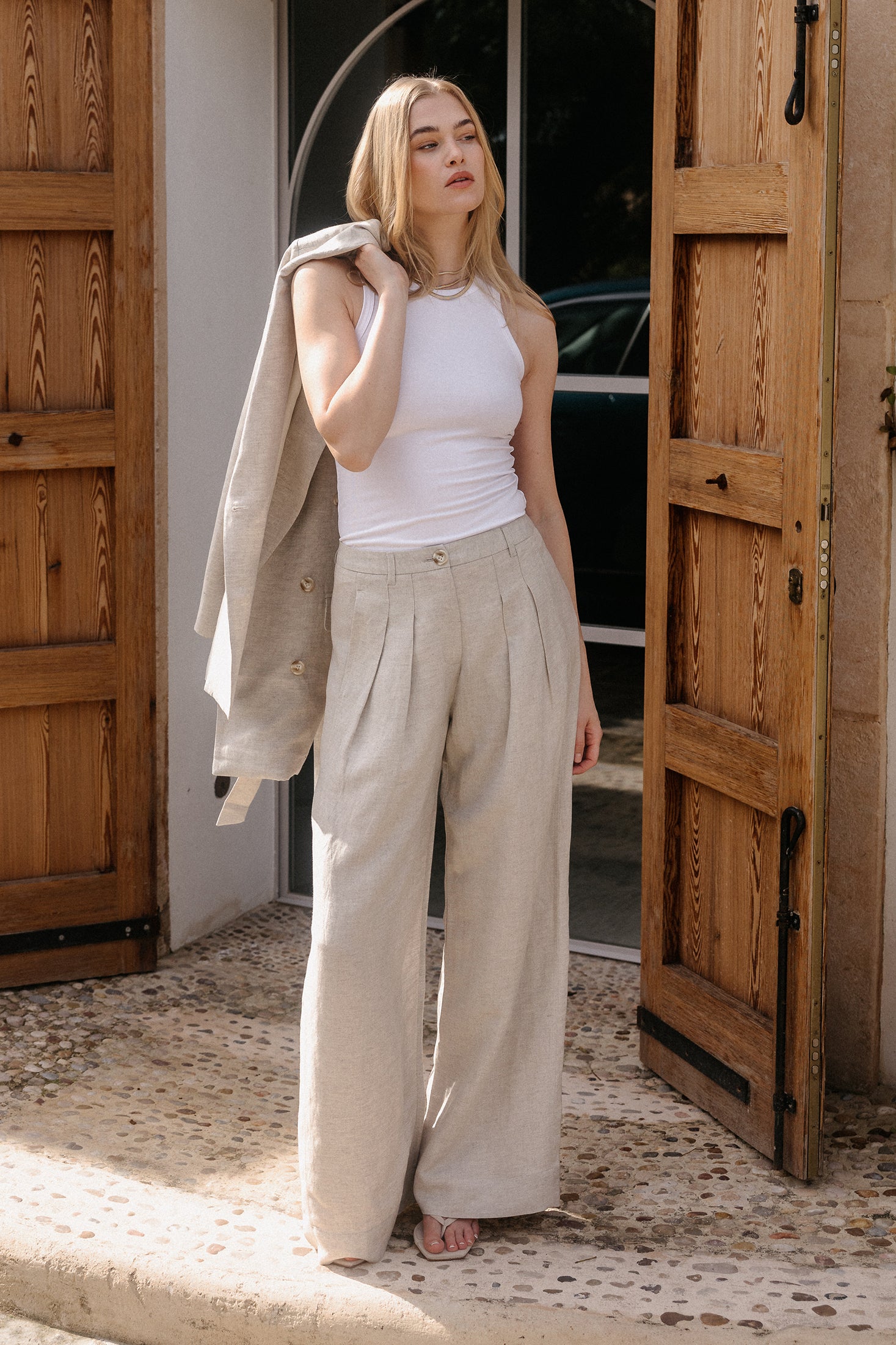 Model in weißem ärmellosem Top und weiter beigefarbener Leinenhose mit passendem Blazer über der Schulter vor offenen Holztüren.