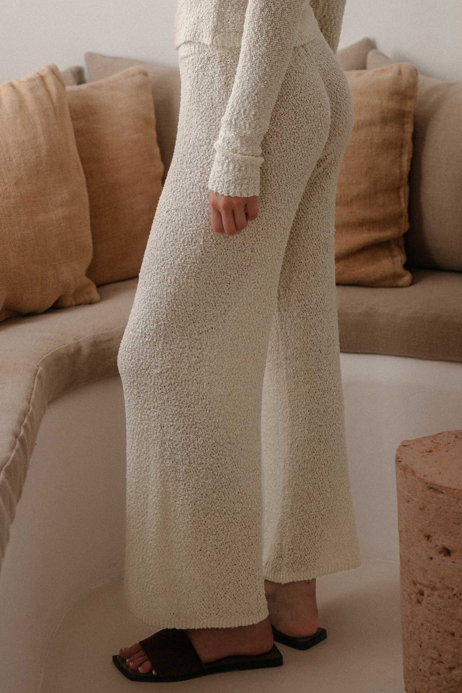 Detailansicht einer cremefarbenen Strickhose in weiter, gerader Passform mit strukturierter Bouclé-Optik. Die Hose wird mit einem passenden langärmligen Strickoberteil kombiniert und in einem hellen, minimalistischen Wohnraum mit beigefarbenen Sitzpolstern und natürlichen Materialien präsentiert.