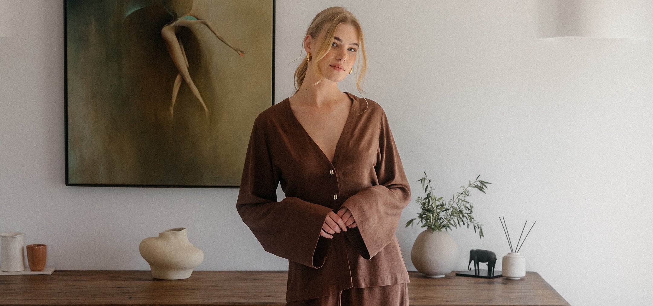 Blonde Frau in braunem Leinen-Zweiteiler vor minimalistischer Einrichtung mit Holzkonsole und neutraler Dekoration.