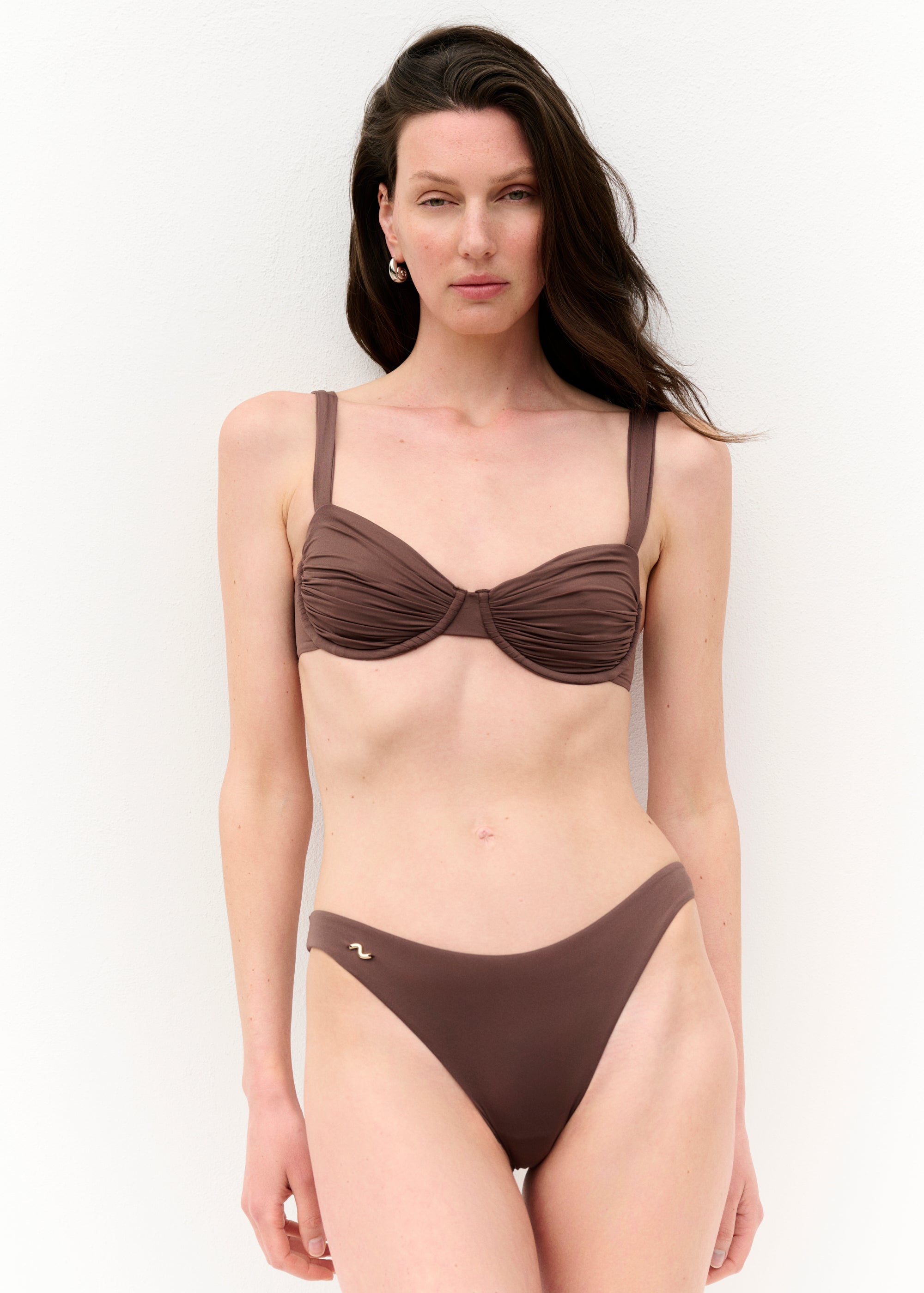 Frau mit langen dunklen Haaren trägt ein braunes Bikini-Set mit gerafftem Bügel-Top und schlichter Bikinihose. Sie steht vor weißem Hintergrund, blickt leicht zur Seite und hält mit einem Arm den anderen. Das Bikini-Top hat breite Träger und goldene Details an der Rückenschließe. Die Hose ist hoch geschnitten und auf einer Seite mit einem kleinen goldenen Logo verziert.