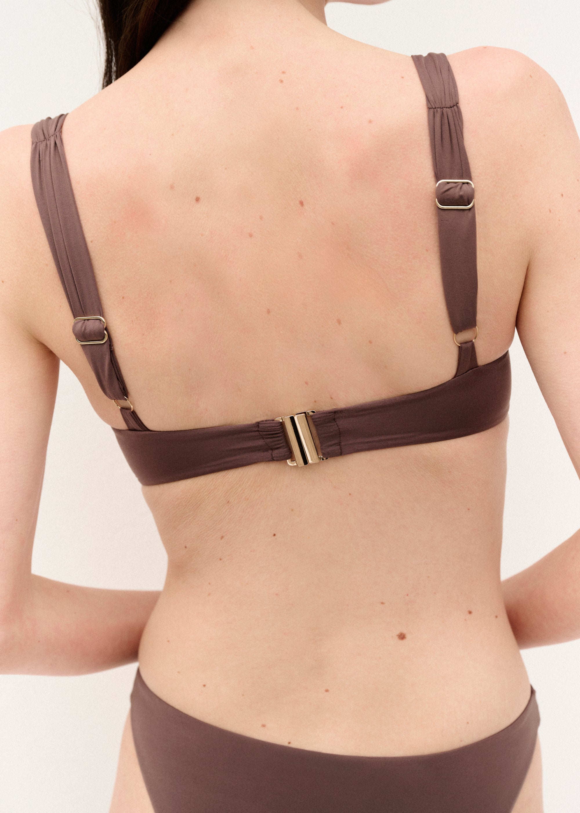 Frau mit langen dunklen Haaren trägt ein braunes Bikini-Set mit gerafftem Bügel-Top und schlichter Bikinihose. Sie steht vor weißem Hintergrund, blickt leicht zur Seite und hält mit einem Arm den anderen. Das Bikini-Top hat breite Träger und goldene Details an der Rückenschließe. Die Hose ist hoch geschnitten und auf einer Seite mit einem kleinen goldenen Logo verziert.