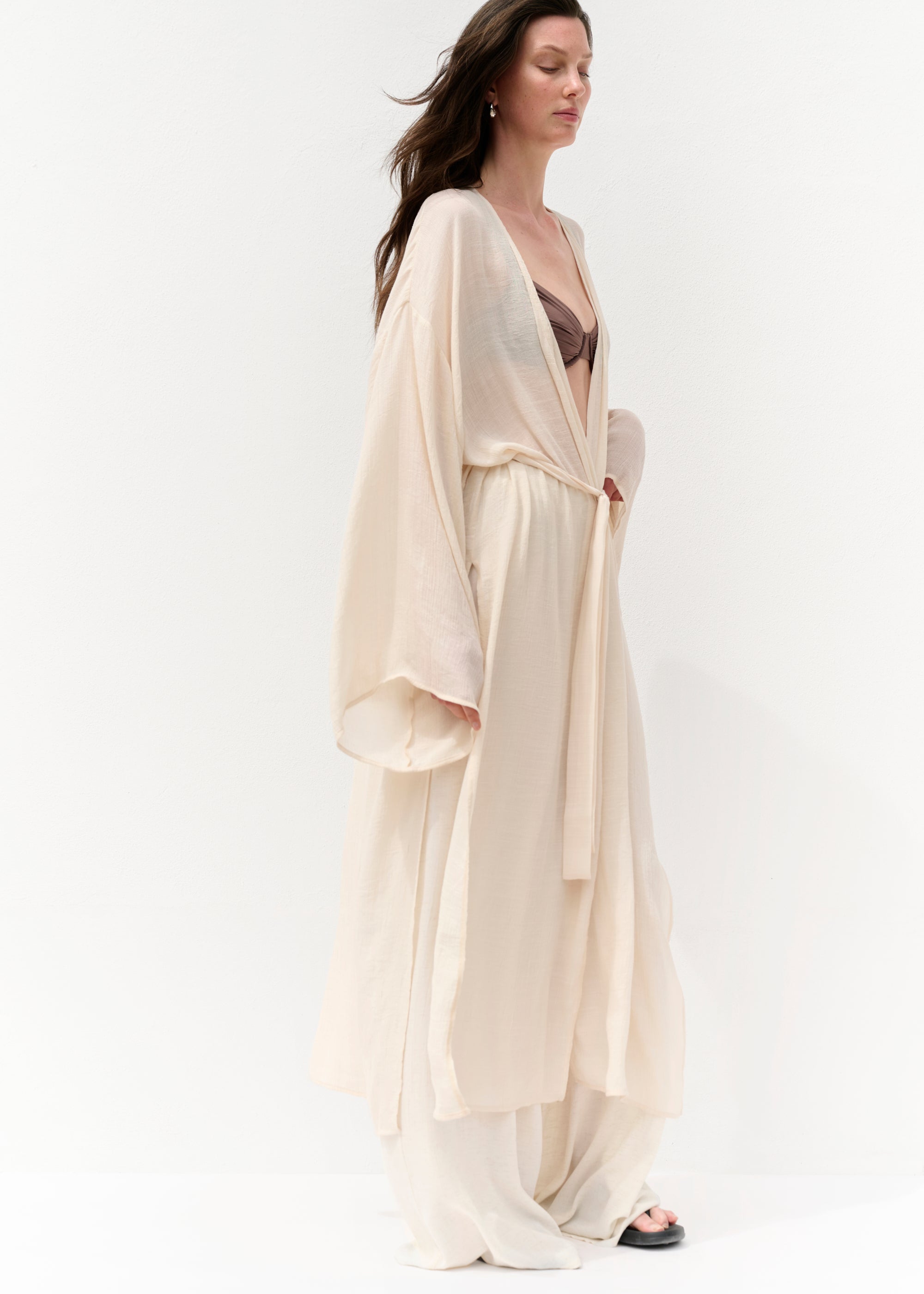 Seitlicher Blick auf eine Frau in einem transparenten, cremefarbenen Cape mit langen, weiten Ärmeln und Bindegürtel. Darunter trägt sie ein braunes Bikini-Oberteil und eine fließende Hose. Ihr Haar weht leicht im Wind, sie trägt schwarze Slides und steht vor einer weißen Wand.