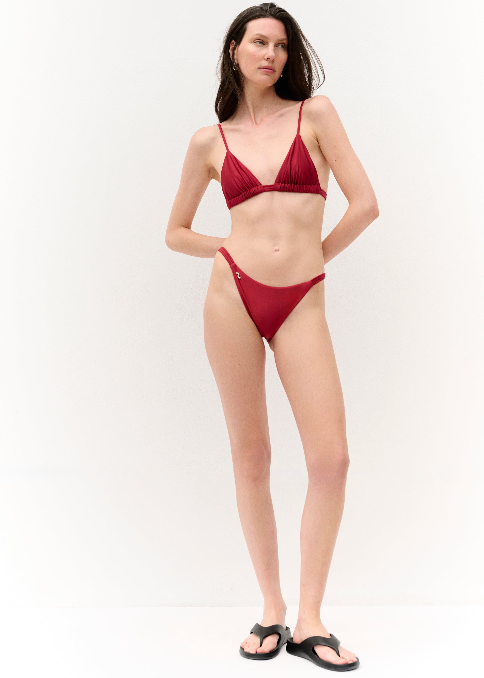 Vollansicht von vorn
Eine Frau steht vor einer weißen Wand und trägt ein dunkelrotes Bikini-Set. Das Oberteil besteht aus zwei dreieckigen Cups mit schmalen Trägern und einer gerafften Unterbrustnaht. Das Höschen ist schmal geschnitten, sitzt hoch auf der Hüfte und hat ein kleines, goldenes Logo-Detail auf der linken Seite.