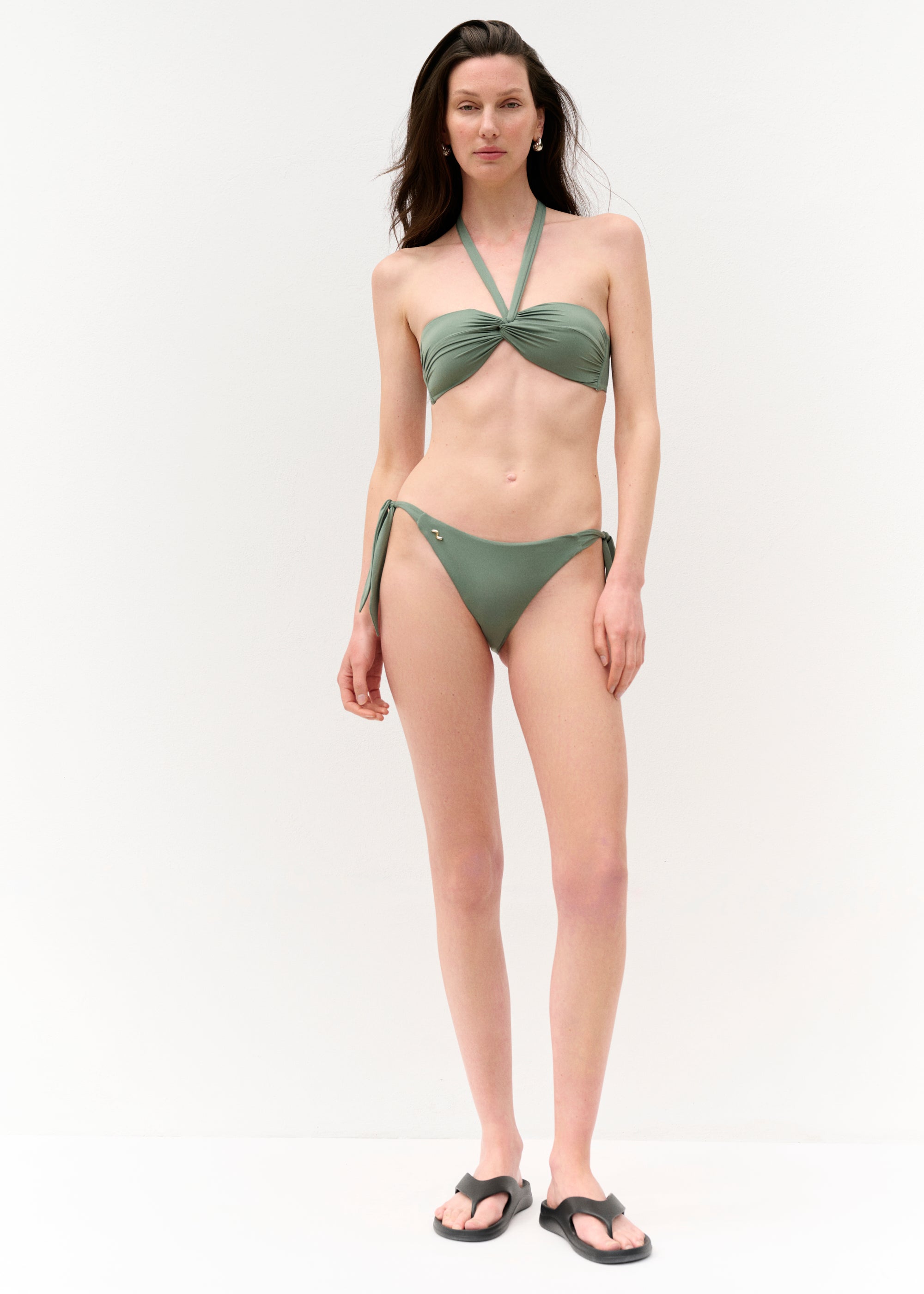 Vollansicht von vorn
Eine Frau steht frontal vor einer weißen Wand und trägt ein olivgrünes Bikini-Set. Das Oberteil ist trägerlos mit einer gedrehten Frontpartie und überkreuzten Neckholder-Trägern. Das Unterteil hat seitliche Bindebänder und ein kleines, goldenes Schmuckdetail auf der linken Seite.