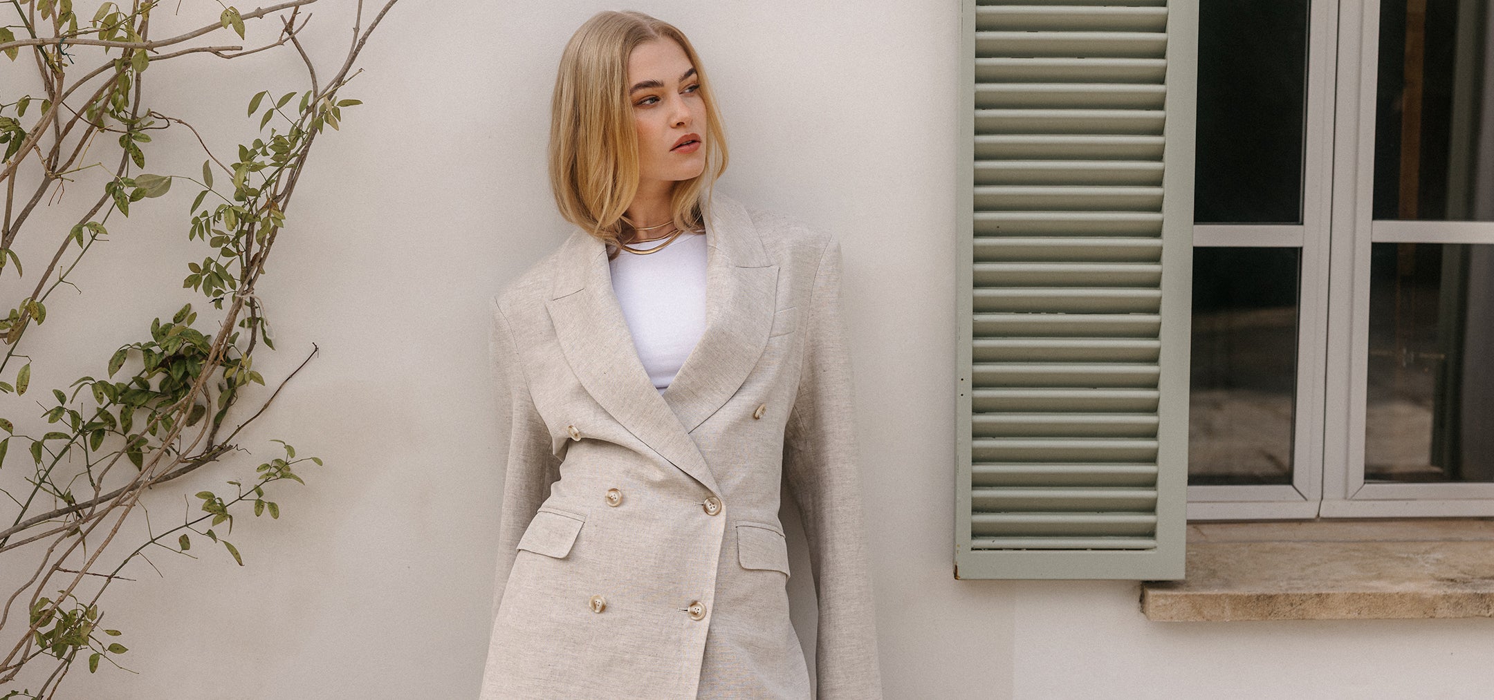 Blonde Frau im hellen doppelreihigen Blazer vor cremefarbener Wand mit Fensterläden und natürlichem Licht.
