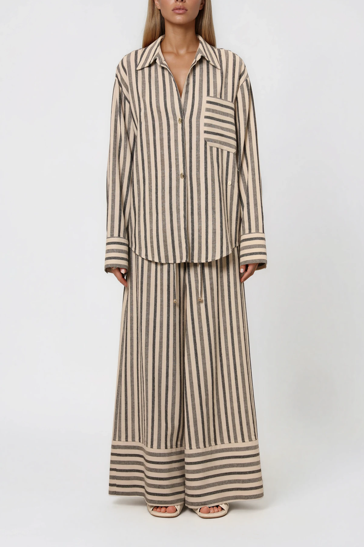 Model trägt eine gestreifte Bluse mit asymmetrischer Knopfleiste und Brusttasche, kombiniert mit einer weit geschnittenen Hose im passenden Streifenmuster.