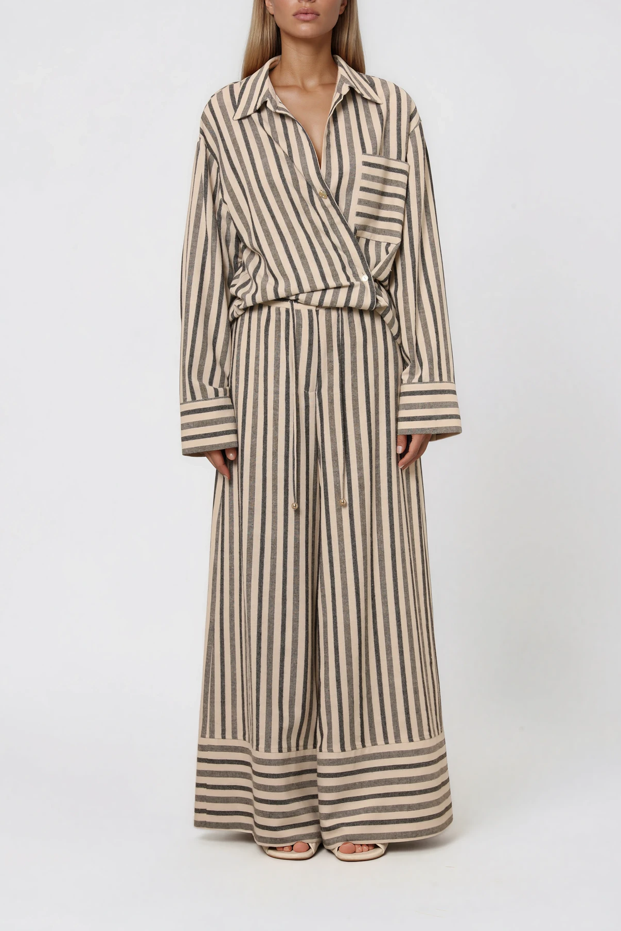 Model trägt eine gestreifte Bluse mit asymmetrischer Knopfleiste und Brusttasche, kombiniert mit einer weit geschnittenen Hose im passenden Streifenmuster.