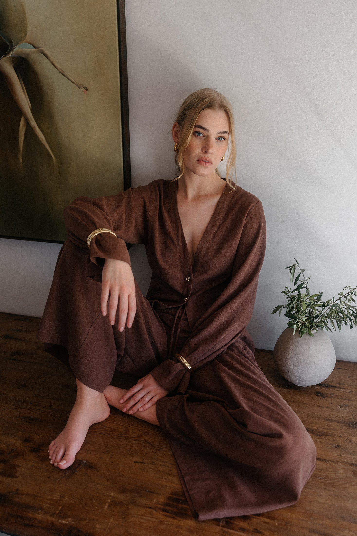 Model sitzt barfuß auf einem Holztisch und trägt ein braunes, locker fallendes Outfit mit langärmliger Bluse mit Knopfleiste und V-Ausschnitt sowie weit geschnittener Hose, kombiniert mit goldenen Armreifen und Ohrringen, vor einer schlichten Wand mit Kunstbild und Vase