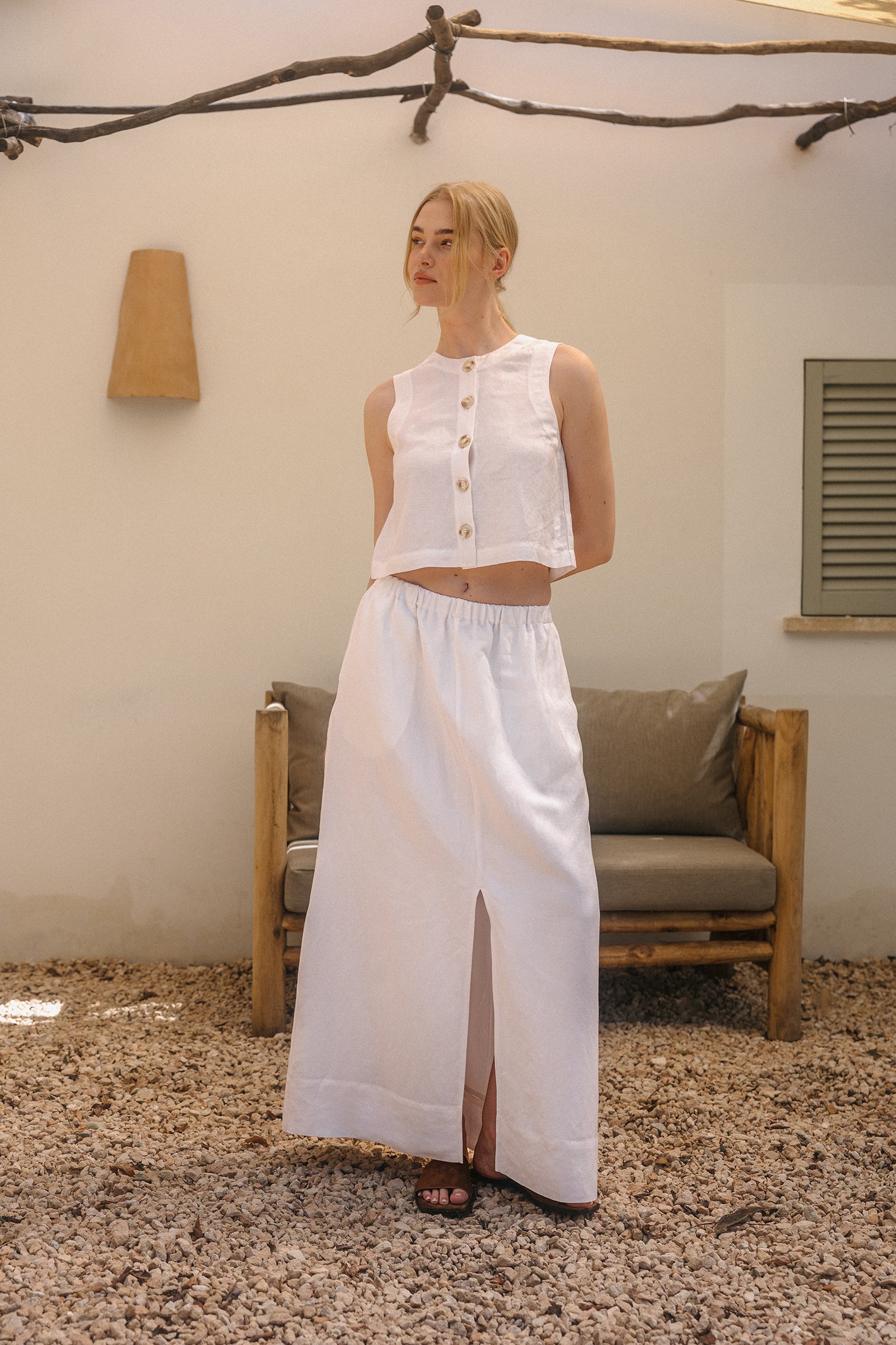 Model trägt ein weißes ärmelloses Crop-Top mit Knopfleiste und Rundhalsausschnitt, kombiniert mit einem langen Rock mit elastischem Bund und Schlitz vorne, steht auf Kiesboden vor einer Sitzbank im Außenbereich