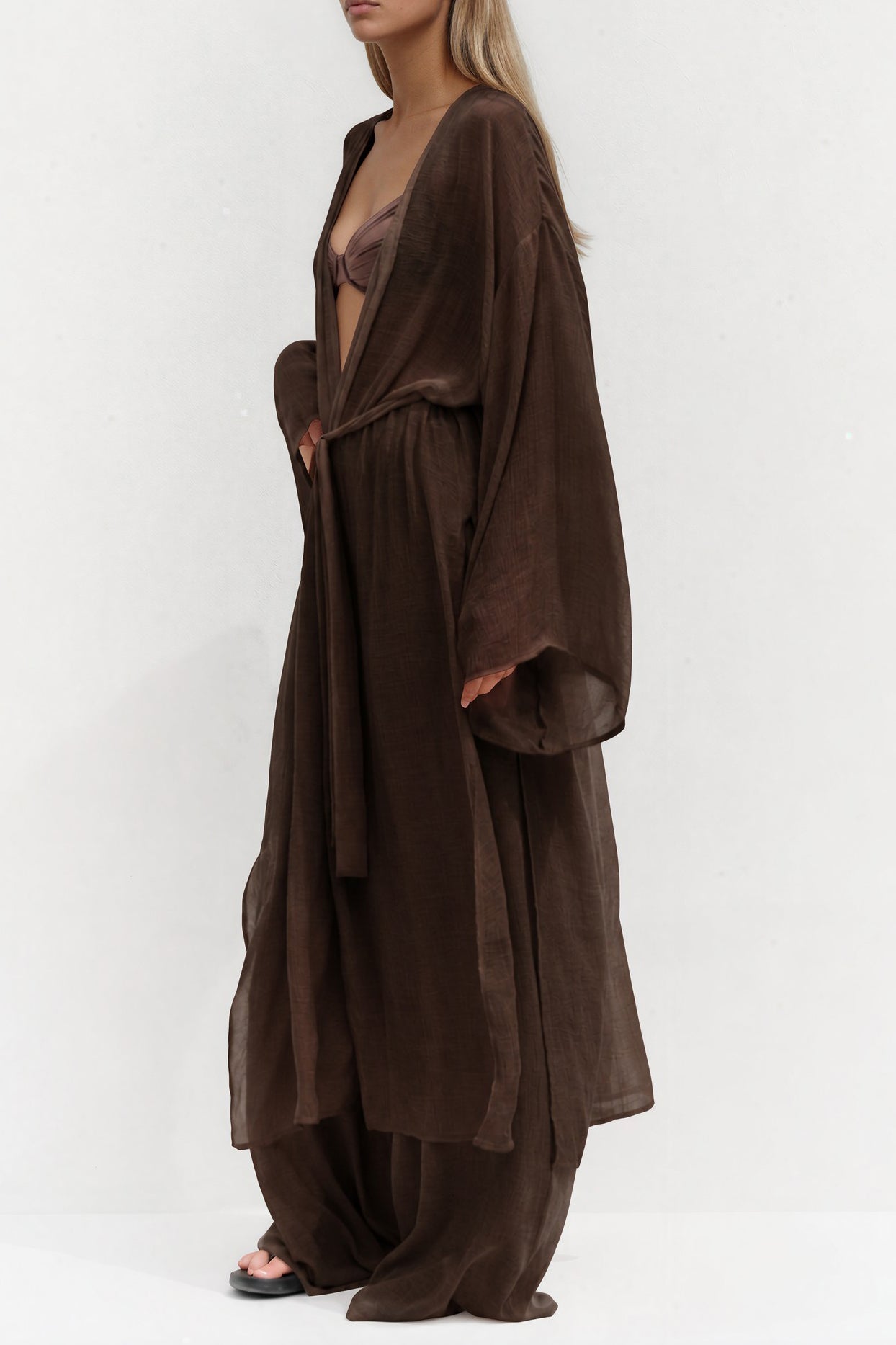 Model trägt einen transparenten dunkelbraunen Kimono mit weitem Schnitt und Bindegürtel über einem braunen Bikini-Top und fließender Hose in passender Farbe.
