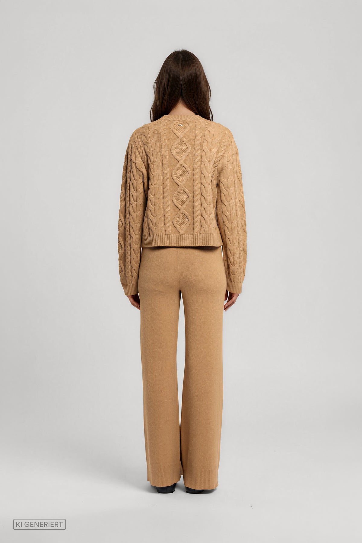 Rückansicht eines Models in einem camelbraunen Strickpullover mit Zopfmuster und Rippbündchen, kombiniert mit einer weit geschnittenen Strickhose.