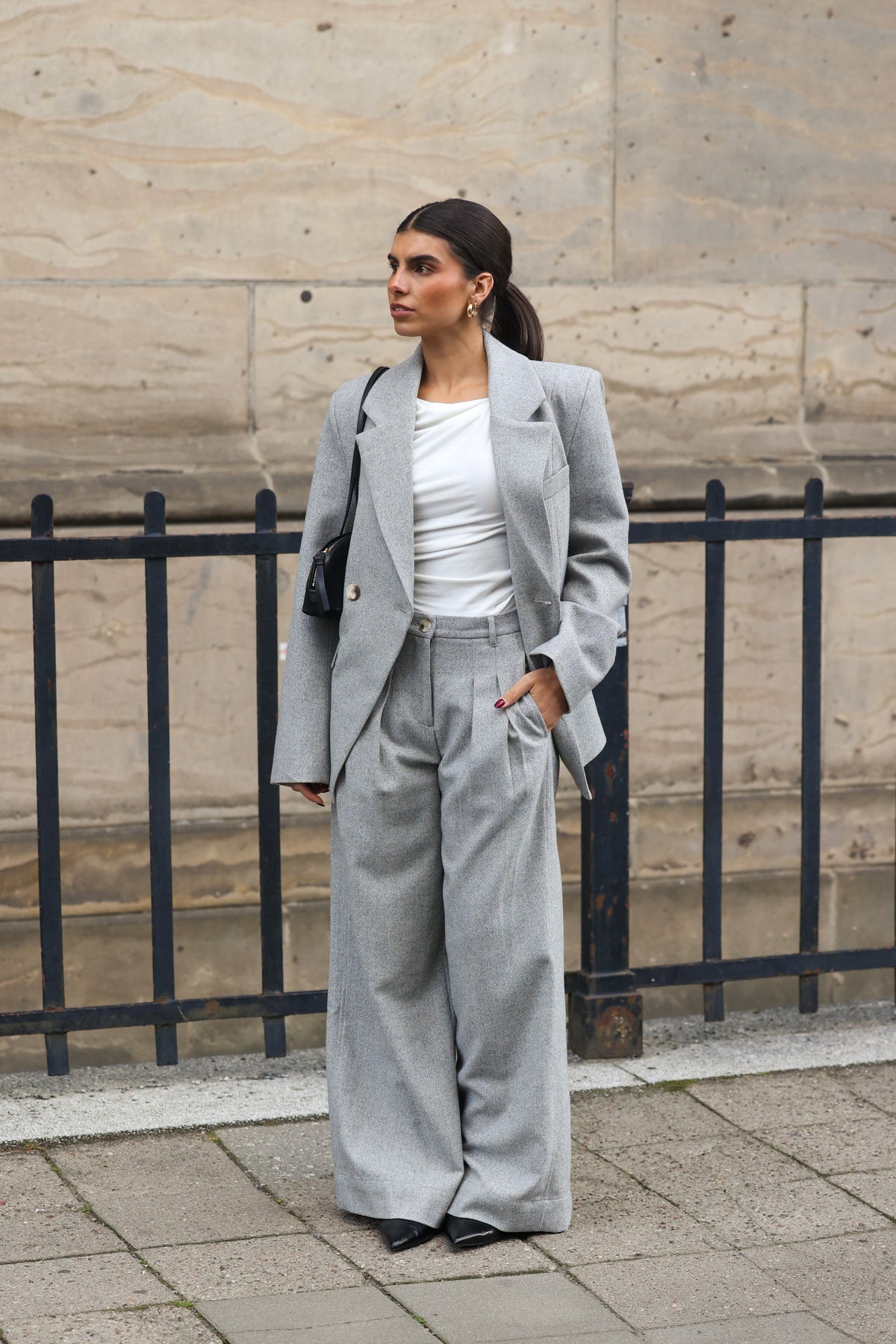 Model trägt die ADA Pants Herringbone Grey von LEZU mit passendem DELIA Blazer und weißem Top, Streetstyle vor einer Steinfassade.