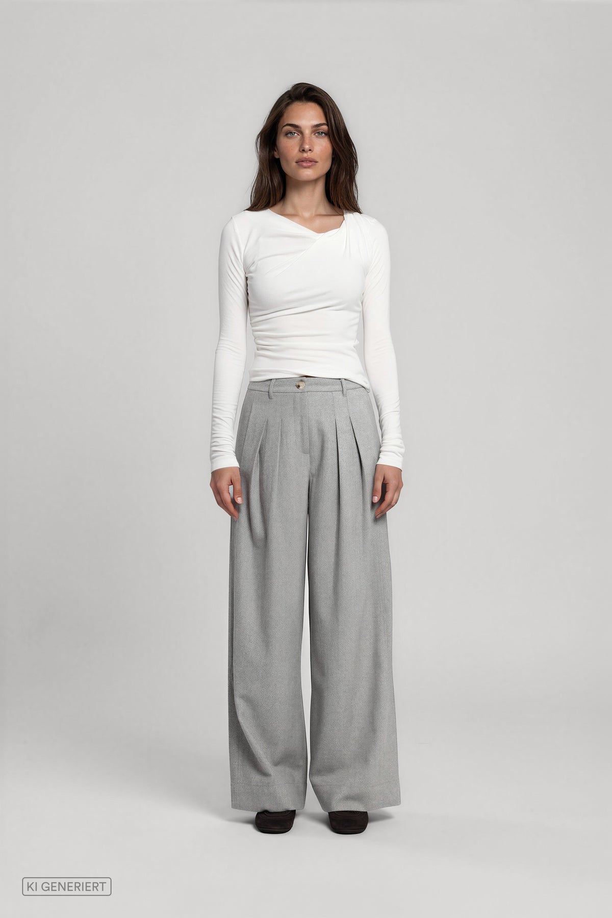 Frontansicht eines Models in einem weißen, figurbetonten Langarmshirt mit asymmetrischer Raffung und einer hellgrauen, weit geschnittenen Stoffhose mit Bundfalten.
