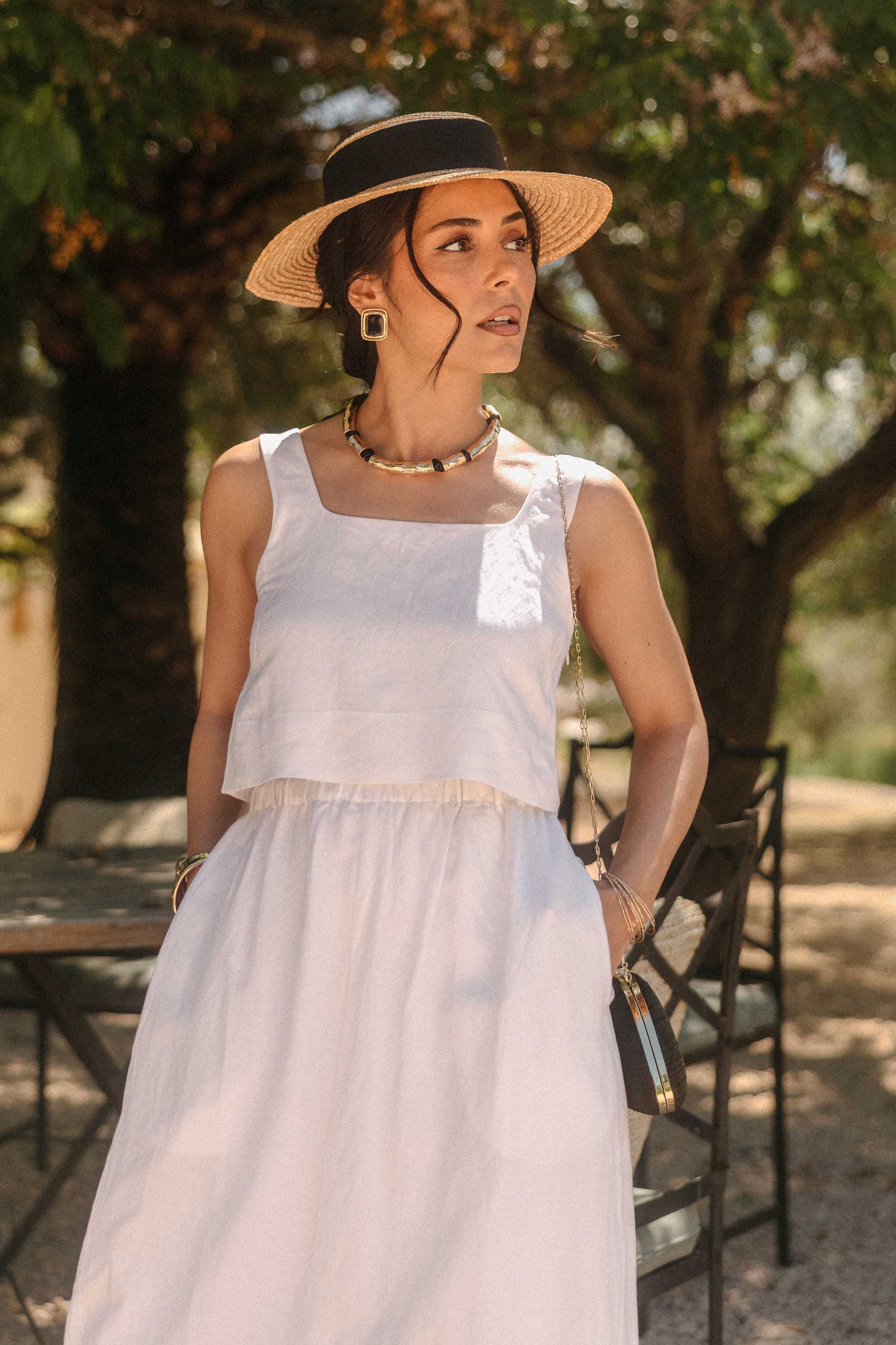Frau in einem weißen Sommerkleid mit locker fallendem Rock steht unter schattenspendenden Bäumen in mediterraner Umgebung. Sie trägt einen flachen Strohhut mit schwarzem Band, auffälligen Schmuck und blickt zur Seite. Das warme Sonnenlicht und die natürliche Kulisse mit Holzmöbeln verleihen der Szene eine elegante, sommerliche Atmosphäre.