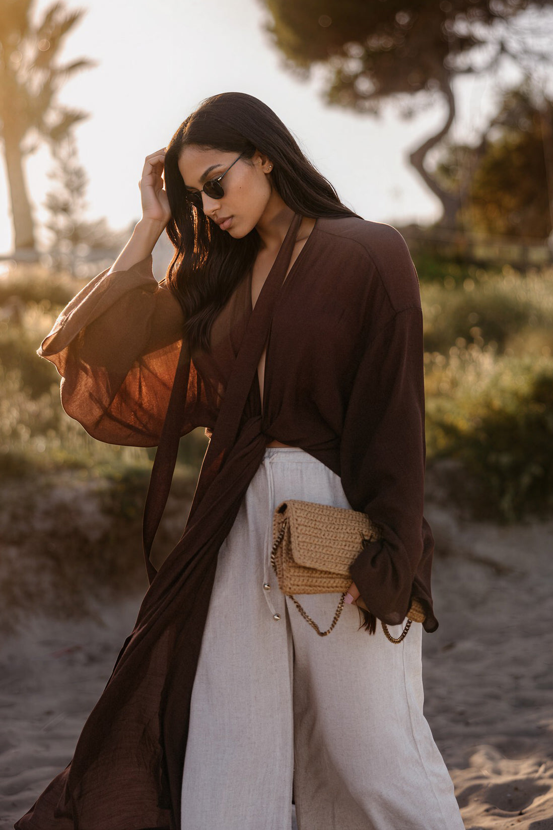 Model in transparentem dunkelbraunem Kimono-Top mit Bindegürtel, weiter heller Leinenhose und geflochtener Basttasche im warmen Abendlicht.