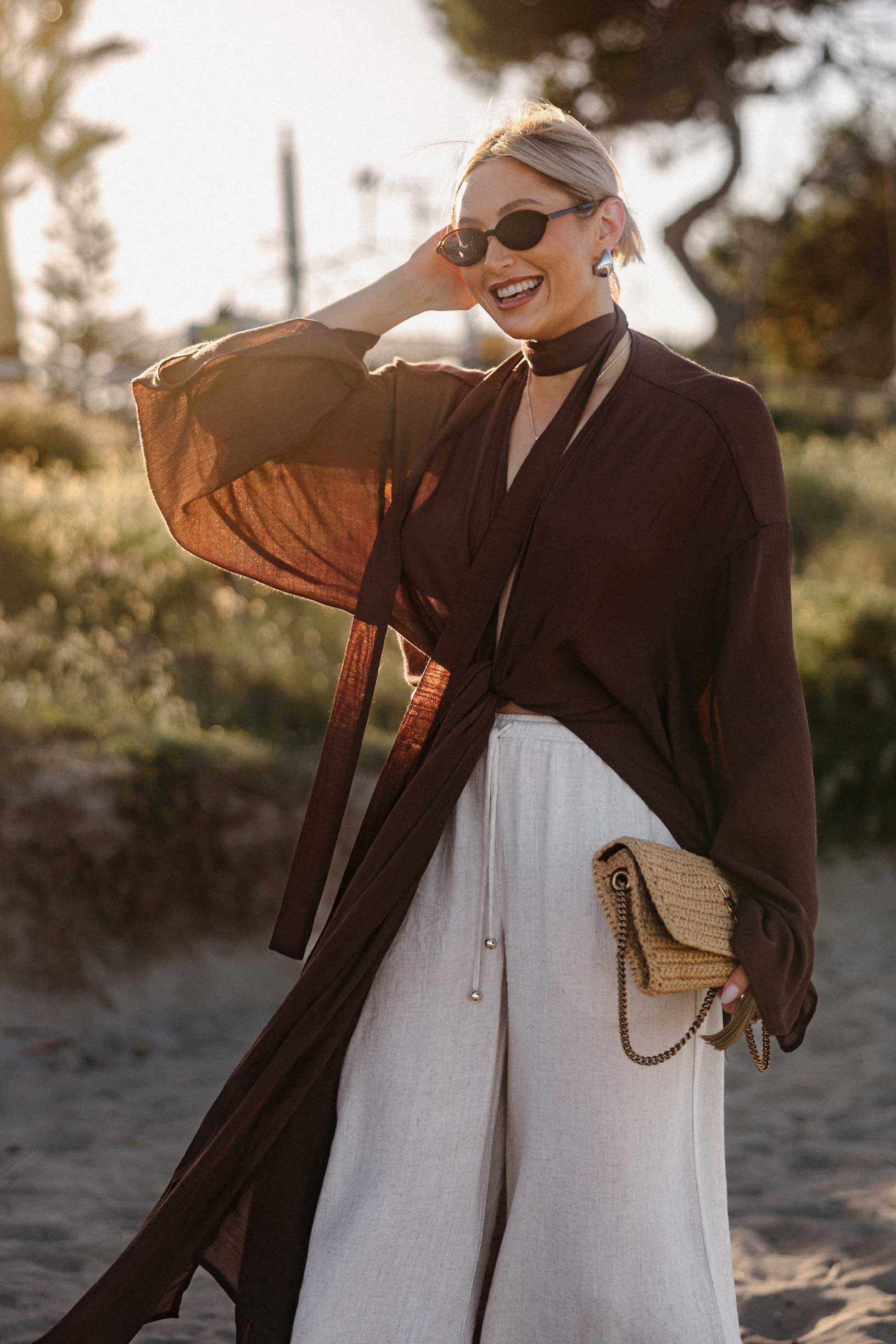 Frau mit Sonnenbrille steht bei goldenem Abendlicht auf einem Sandweg am Strand. Sie trägt eine weite cremefarbene Hose und einen dunkelbraunen, luftigen Kimono mit Bindegürtel und hält eine geflochtene Clutch in der Hand. Palmen und mediterrane Vegetation bilden den unscharfen Hintergrund.