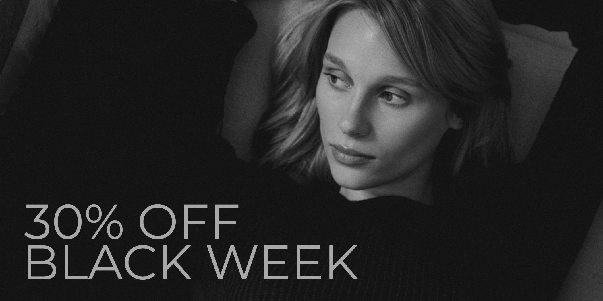 Schwarz-weißes Bannerfoto einer jungen Frau mit offenen blonden Haaren in entspannter Pose, die auf dem Rücken liegt und zur Seite schaut. Sie trägt ein schwarzes geripptes Oberteil. Links im Bild steht in heller Schrift: „30% OFF BLACK WEEK“.
