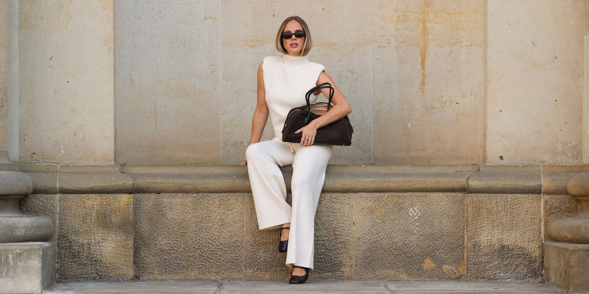 Frau in einem cremeweißen Strick-Look aus ärmellosem Top und weiter Hose sitzt vor einer Natursteinwand und trägt eine schwarze Handtasche und Sonnenbrille.