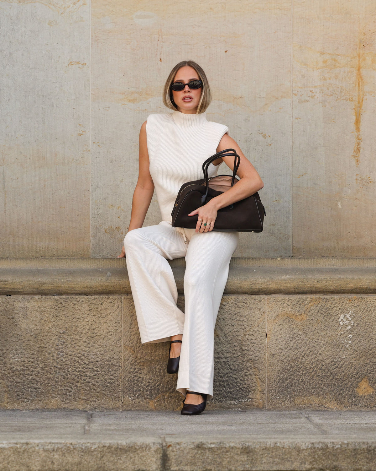 Frau in einem cremeweißen Strick-Look aus ärmellosem Top und weiter Hose sitzt vor einer Natursteinwand und trägt eine schwarze Handtasche und Sonnenbrille.