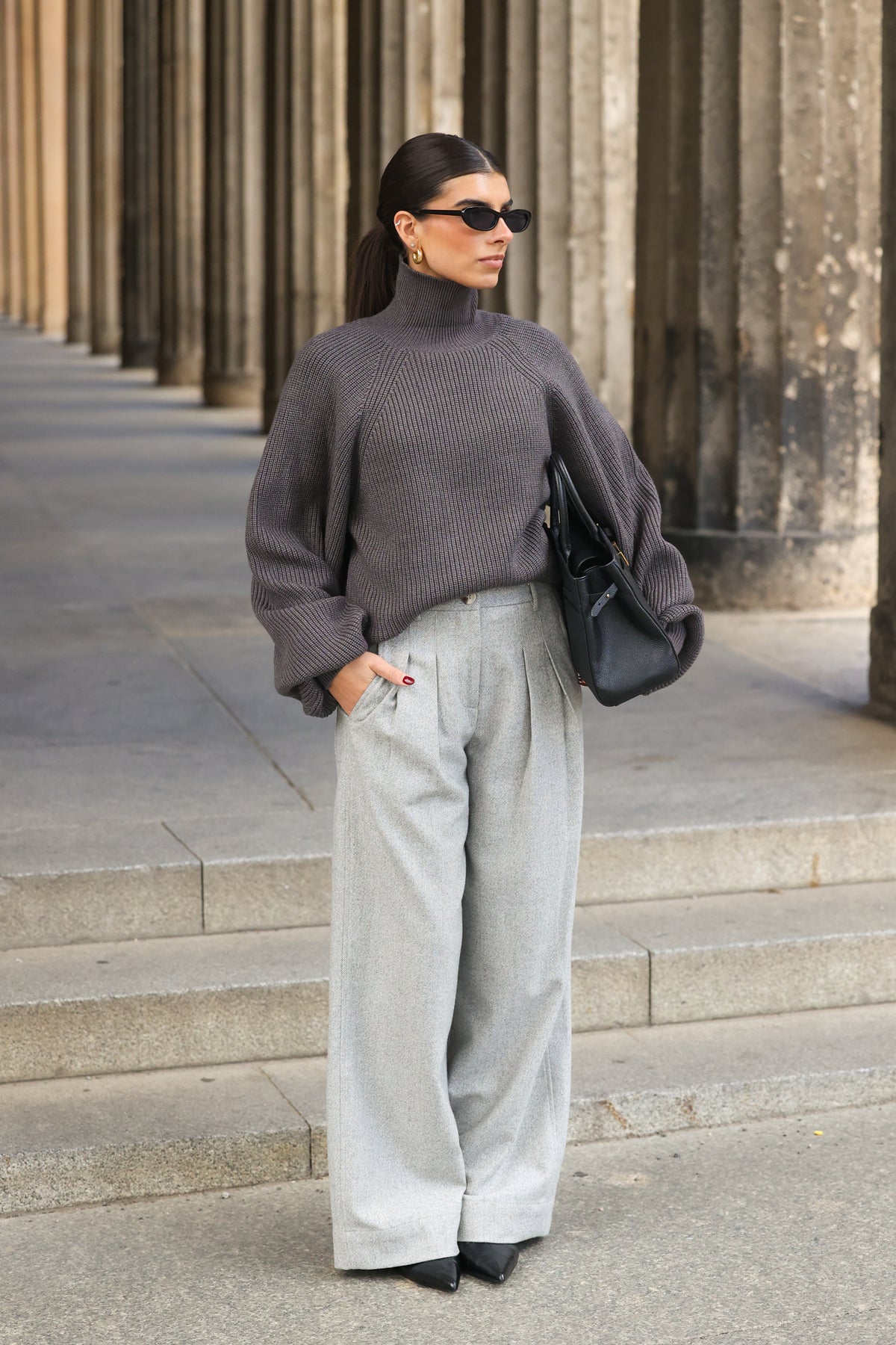 Model in den ADA Pants Herringbone Grey von LEZU mit grauem Rollkragenpullover, Sonnenbrille und schwarzer Tasche vor einer Säulenreihe.