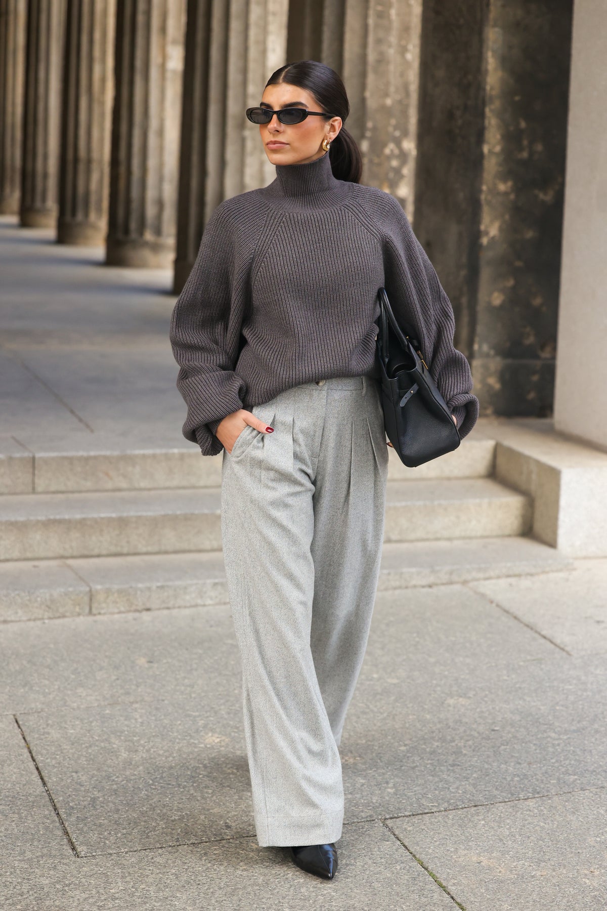 Model läuft in den ADA Pants Herringbone Grey mit weitem Bein und Bundfalten, kombiniert mit grauem Rollkragenpullover, vor einer Säulenreihe.