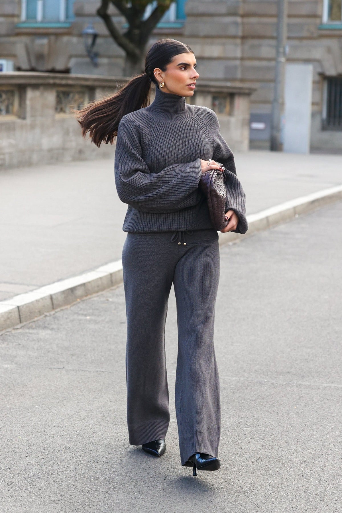 Frau mit dunklem, hohem Pferdeschwanz und goldenen Ohrringen trägt einen grauen Rollkragenpullover aus Strick mit passender weiter Strickhose, schwarze Pumps und eine dunkle Clutch.