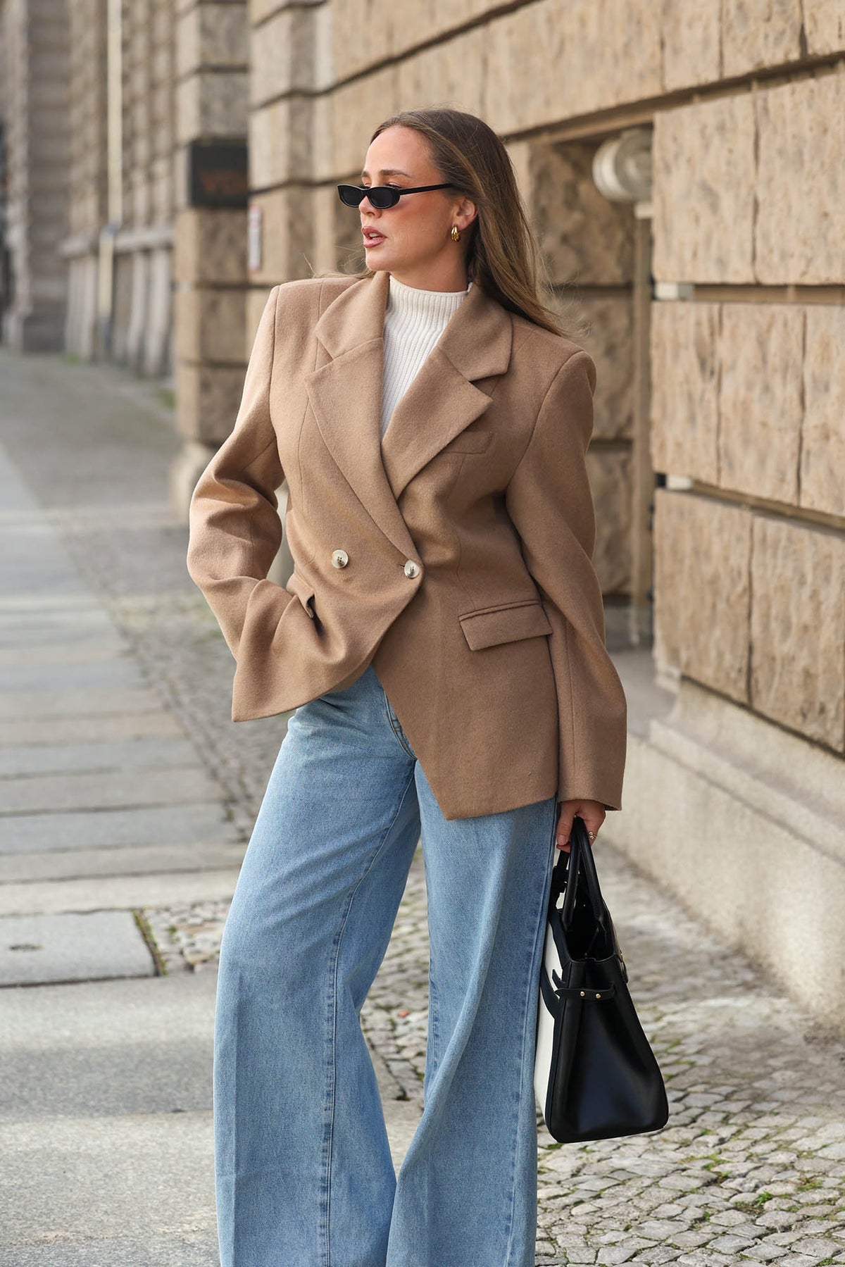 Frau trägt einen beigefarbenen, taillierten Doppelreiher-Blazer über einem weißen Stricktop, kombiniert mit hellblauer Wide-Leg-Jeans, schwarzer Handtasche und Sonnenbrille.