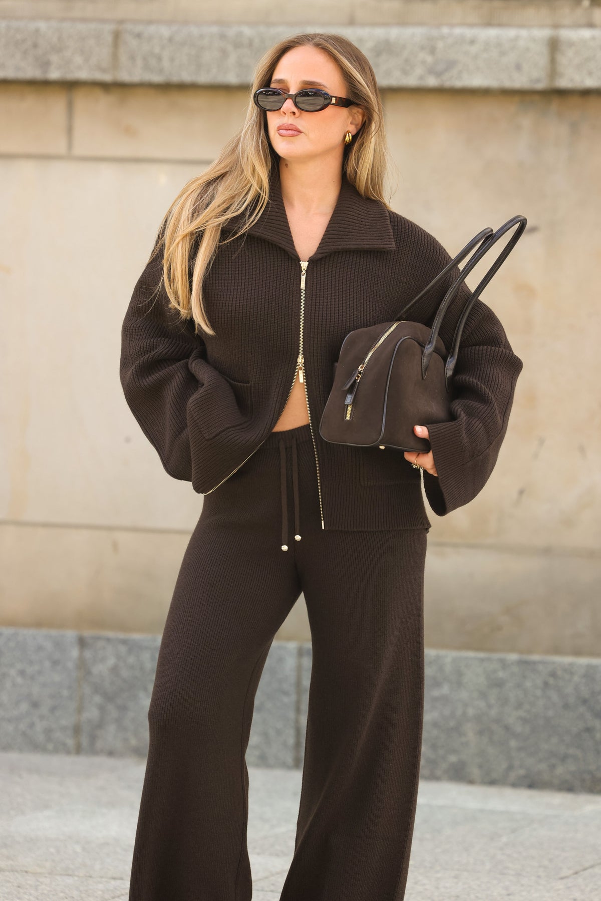 Frau in dunkelbraunem Strick-Cardigan mit Zwei-Wege-Reißverschluss, breitem Kragen und weiter Strickhose, kombiniert mit schwarzer Sonnenbrille und dunkelbrauner Tasche.