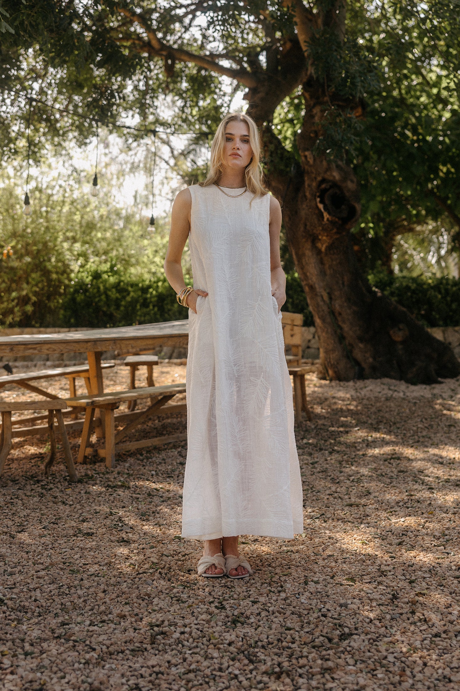 Model steht im Freien unter einem großen Baum und trägt ein langes, ärmelloses weißes Kleid mit geradem Schnitt und seitlichen Taschen, kombiniert mit Sandalen und goldenen Armreifen