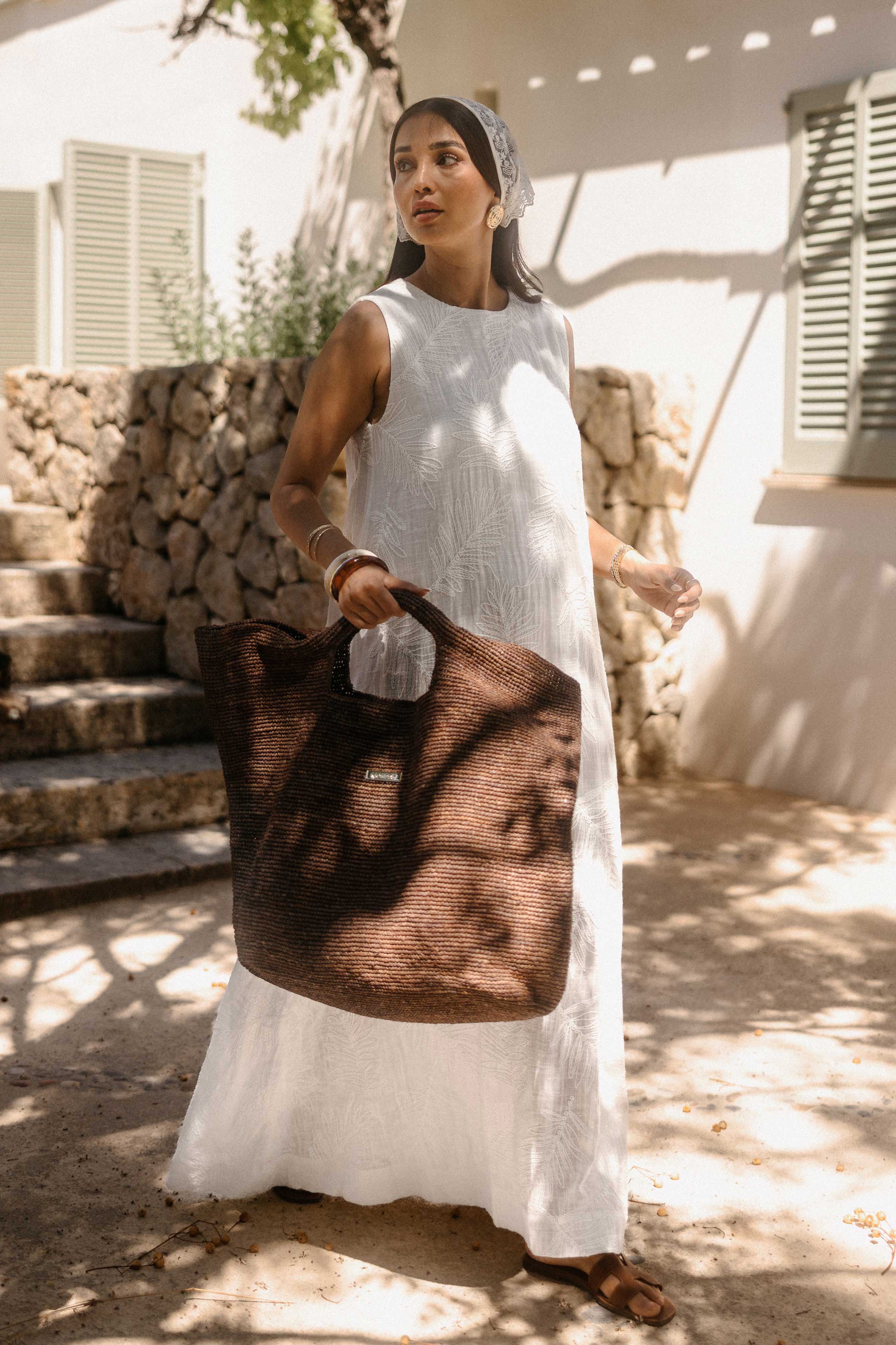 Frau in einem weißen, ärmellosen Minikleid mit eingewebtem Blattmuster steht im Schatten mediterraner Bäume vor einem Landhaus. Sie trägt goldene Ohrringe, eine kleine braune Schultertasche und blickt zur Seite. Das warme Sonnenlicht erzeugt ein natürliches Schattenspiel auf Kleid und Umgebung.
